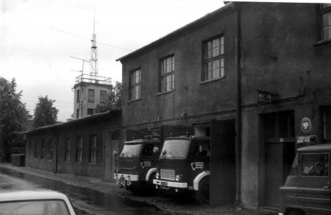 Zdjęcie czarno białe dwupiętrowego budynku zawodowej straży pożarnej w Lublińcu w 1952 roku. Widok ściany frontowej z dwoma bramami wyjazdowymi oraz wejściem głównym do komendy. W otwartych bramach stoją samochody gaśnicze. W tle trzypiętrowa wieża strażacka z masztem radiowym.