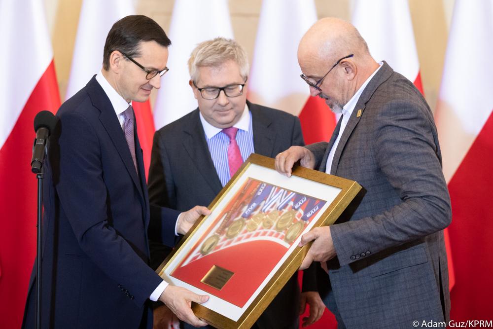 Przedstawiciel PZPS wręcza premierowi oprawione w ramę medale, a z tyłu stoi Ryszard Czarnecki.