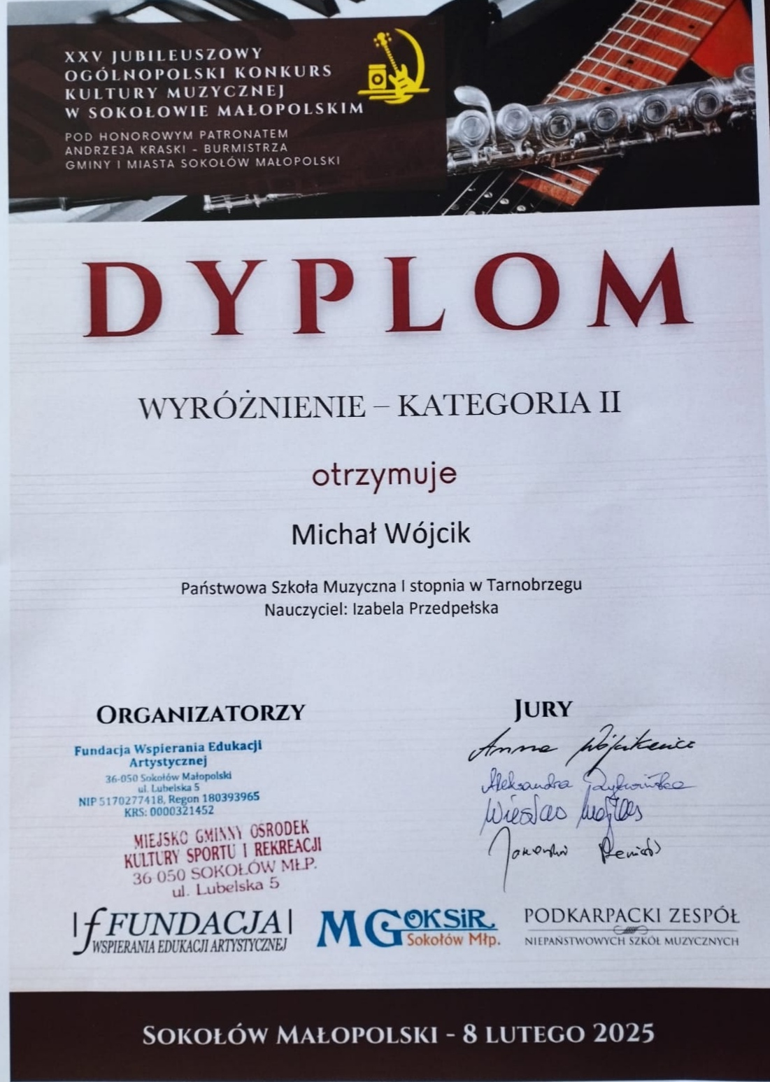 Dyplom wyróżnienia. Na górze znajduje się nazwisko patrona w brązowej ramce, a w tle zdjęcie przedstawiające fragmenty fortepianu, gitary oraz fleta poprzecznego. Po środku nazwisko otrzymującego wyróżnienie oraz nauczyciela. Na dole po lewej stronie organizatorzy, a po prawej podpisy jury. Tekst w kolorze czerwonym i czarnym.