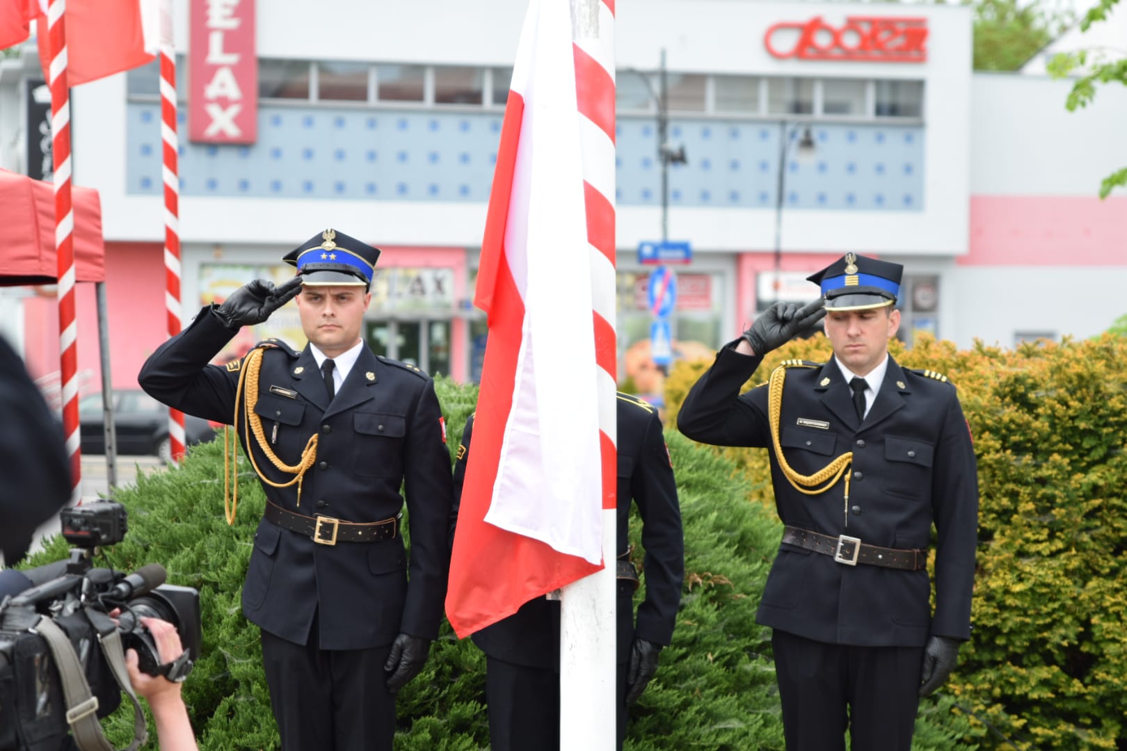 Dwóch strażaków salutuje przy maszcie na którym widnieje flaga RP