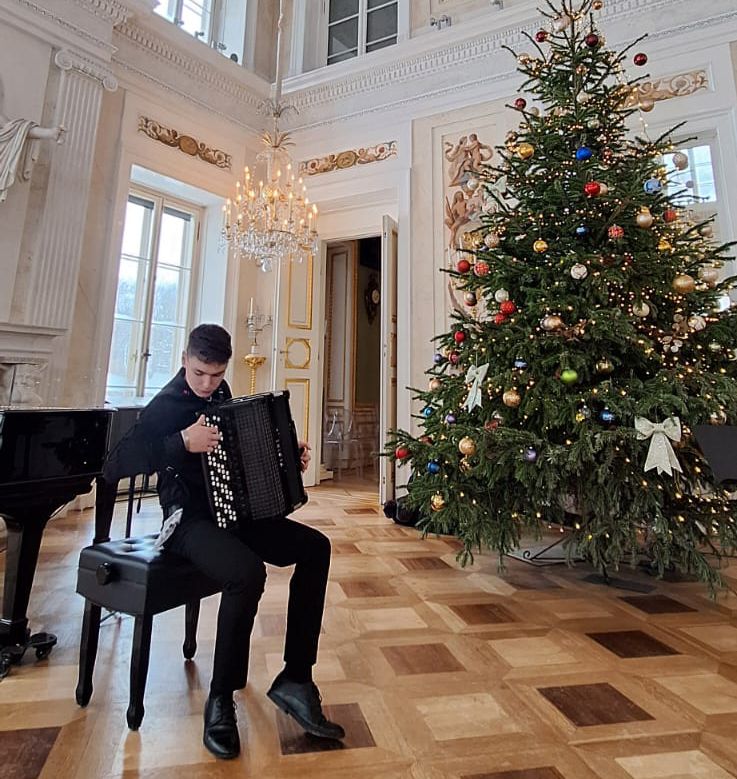 Zdjęcie - Mateusz Neckar na próbie przed recitalem w Pałacu Łazienkowskim. Mateusz gra na akordeonie w sali pałacowej, w tle choinka.