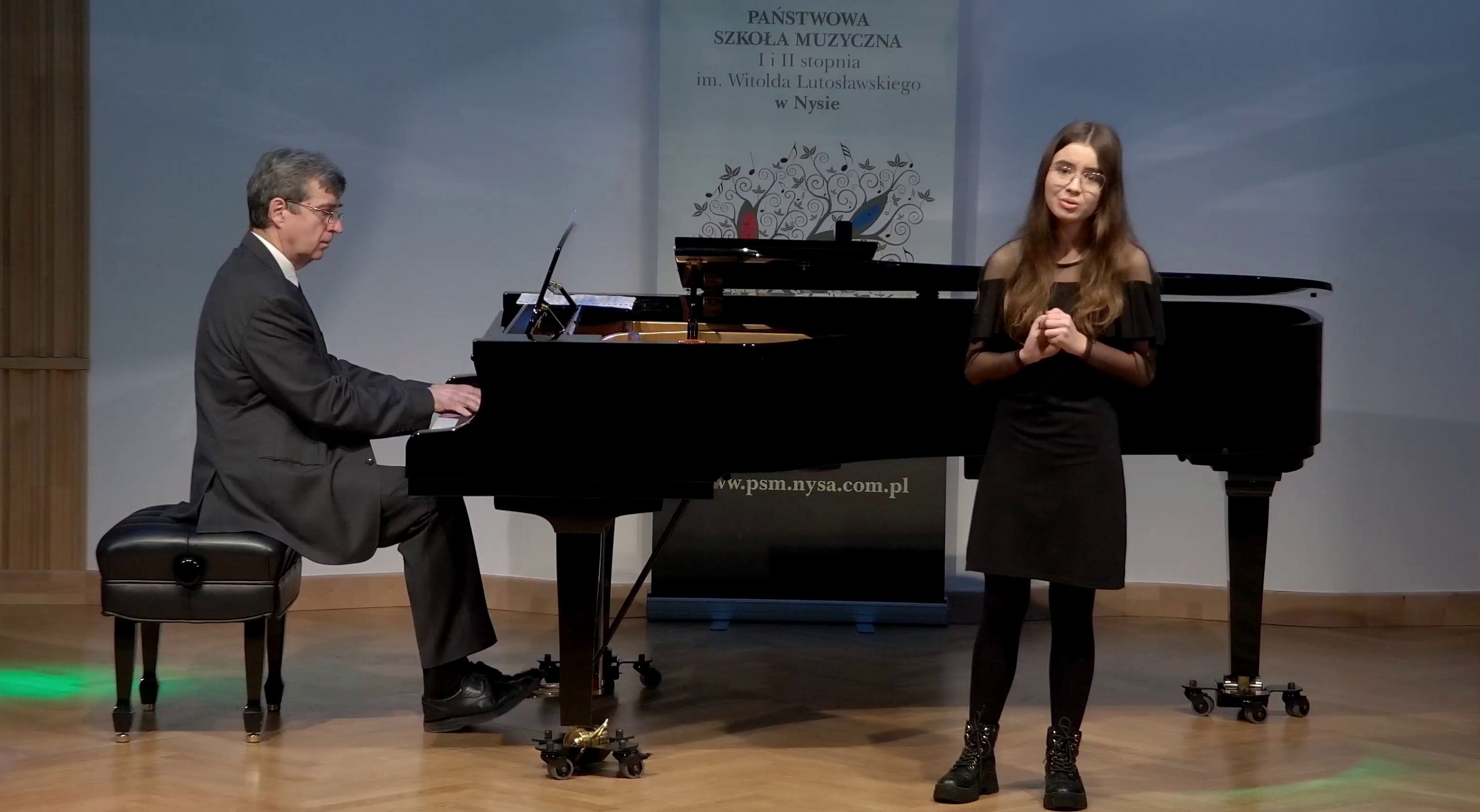 Zdjęcie. Estrada Sali Koncertowej PSM w Nysie. Występ młodej śpiewaczki i pianisty grającego na dużym czarnym fortepianie. Z tyłu w tle baner PSM Nysa.