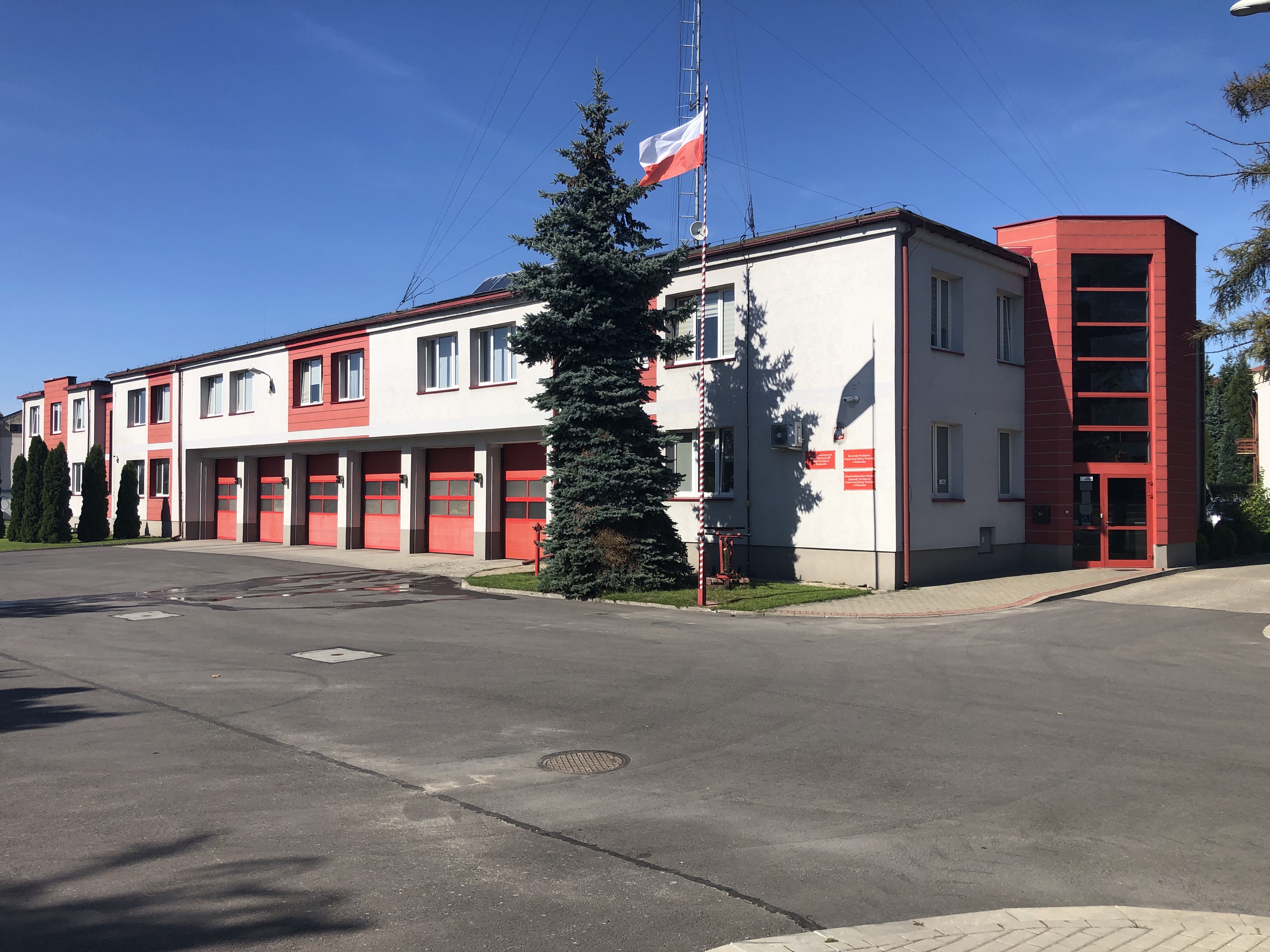 Budynek komendy koloru szarego z czerwonymi elementami i bramami. Przed budynkiem maszt z flagą państwową. 