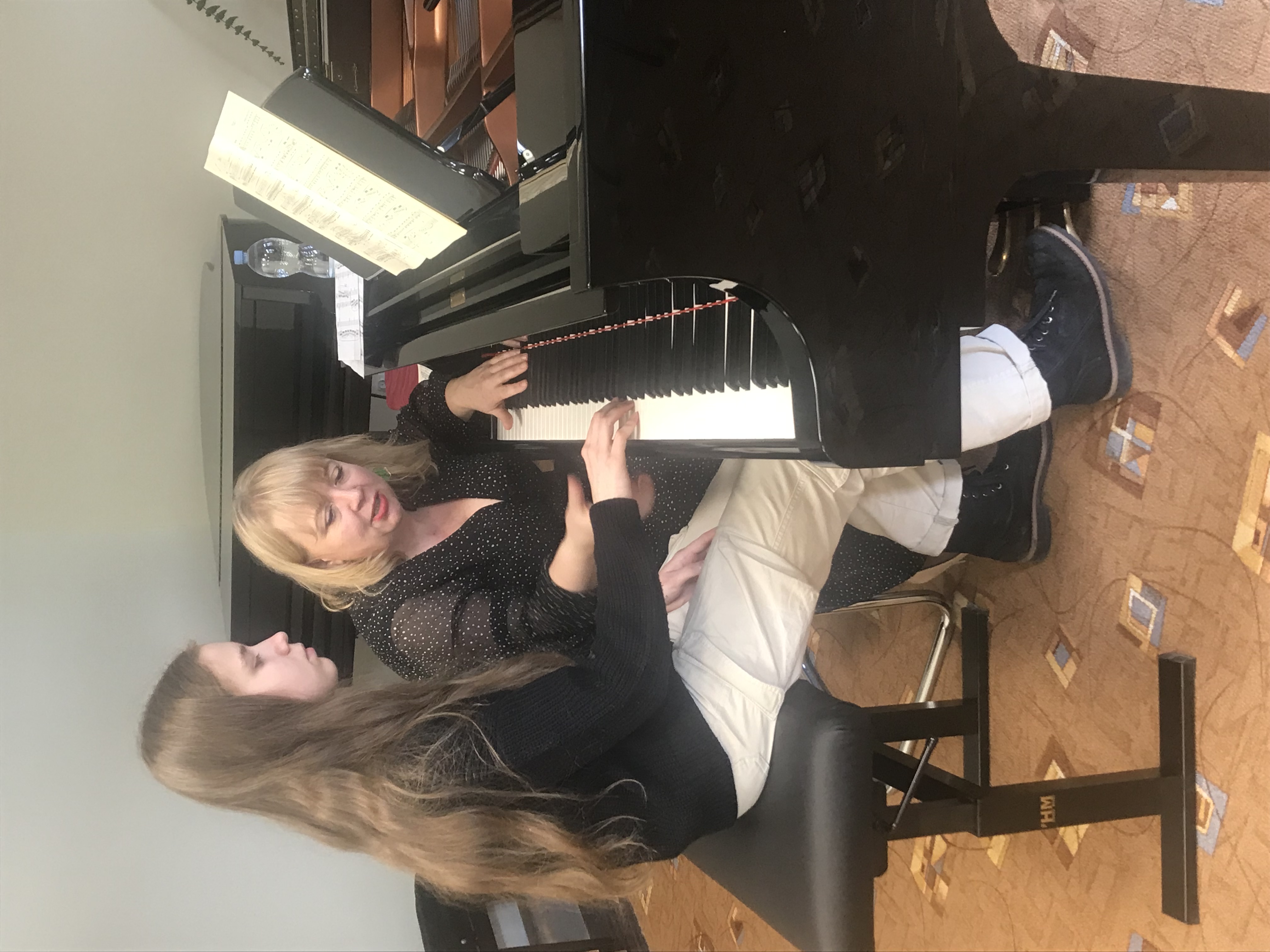 Uczennica siedząca przy fortepianie, obok niej nauczycielka. Z tyłu widoczny jest drugi fortepian.