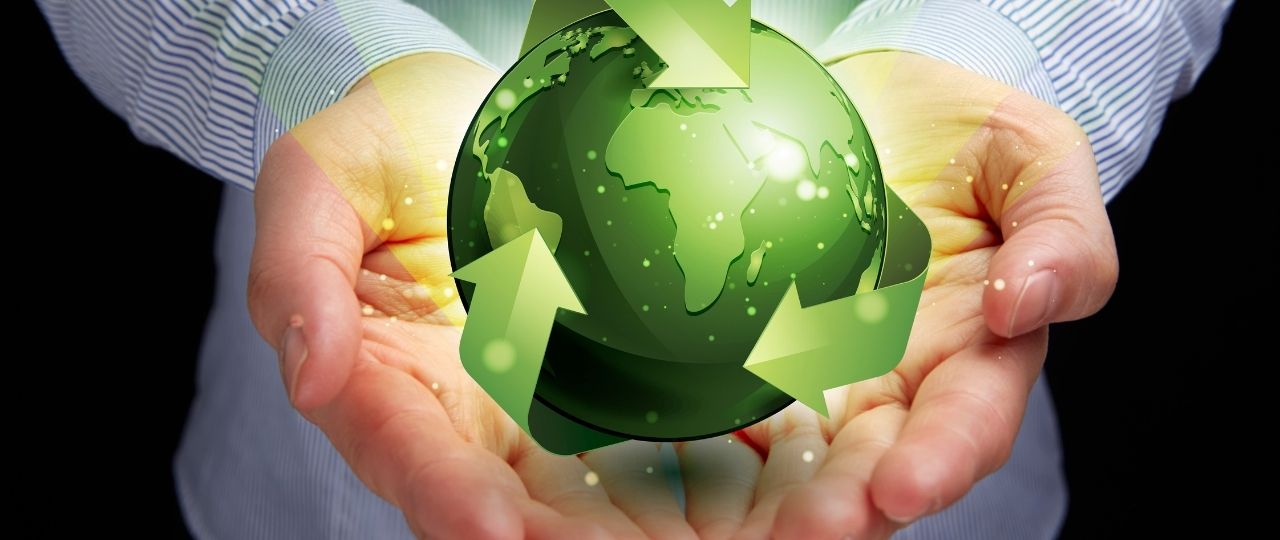 Na ciemnym tle widoczne są dwie dłonie, które delikatnie trzymają zieloną kulę ziemską. Kula przedstawia kontynenty i jest otoczona strzałkami, które tworzą okrąg wokół niej. Strzałki symbolizują ruch lub recykling. Zielony kolor kuli i strzałek świeci, co sprawia, że są dobrze widoczne na tle. Obraz może kojarzyć się z ochroną środowiska, troską o planetę i zrównoważonym rozwojem.