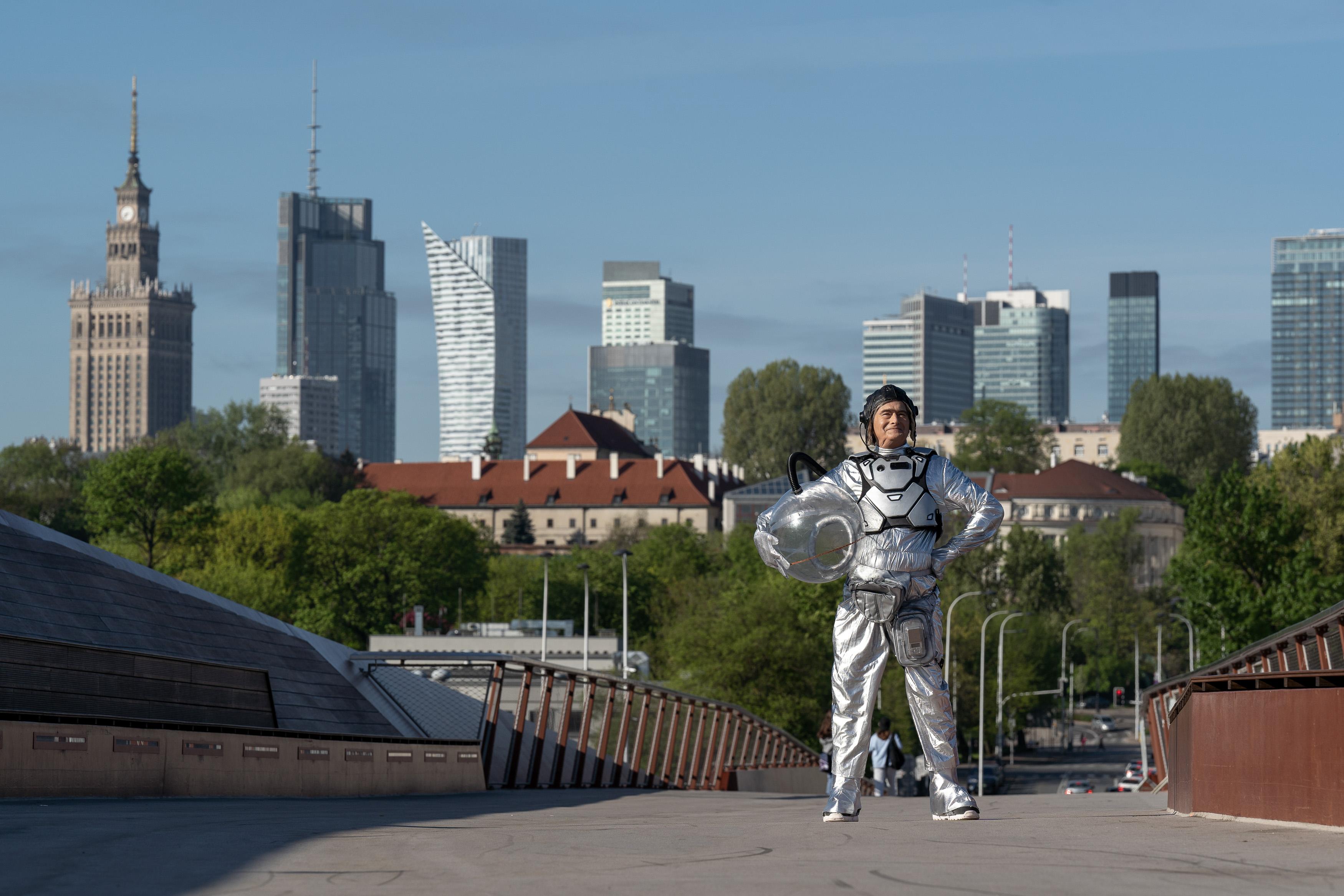 Uśmiechnięty mężczyzna w stroju astronauty stoi na kładce, w tle panorama wieżowców Warszawy