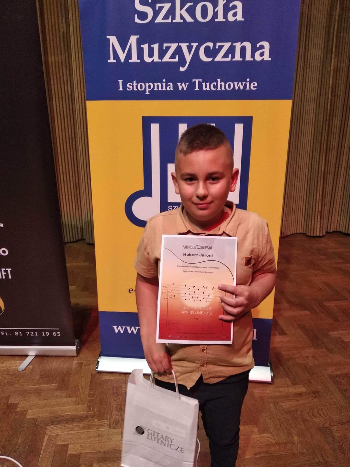 Uczeń trzymający dyplom laureata oraz nagrodę w białej torebce. Z tyłu biały napis "Szkoła Muzyczna I stopnia w Tuchowie" na niebieskim tle.