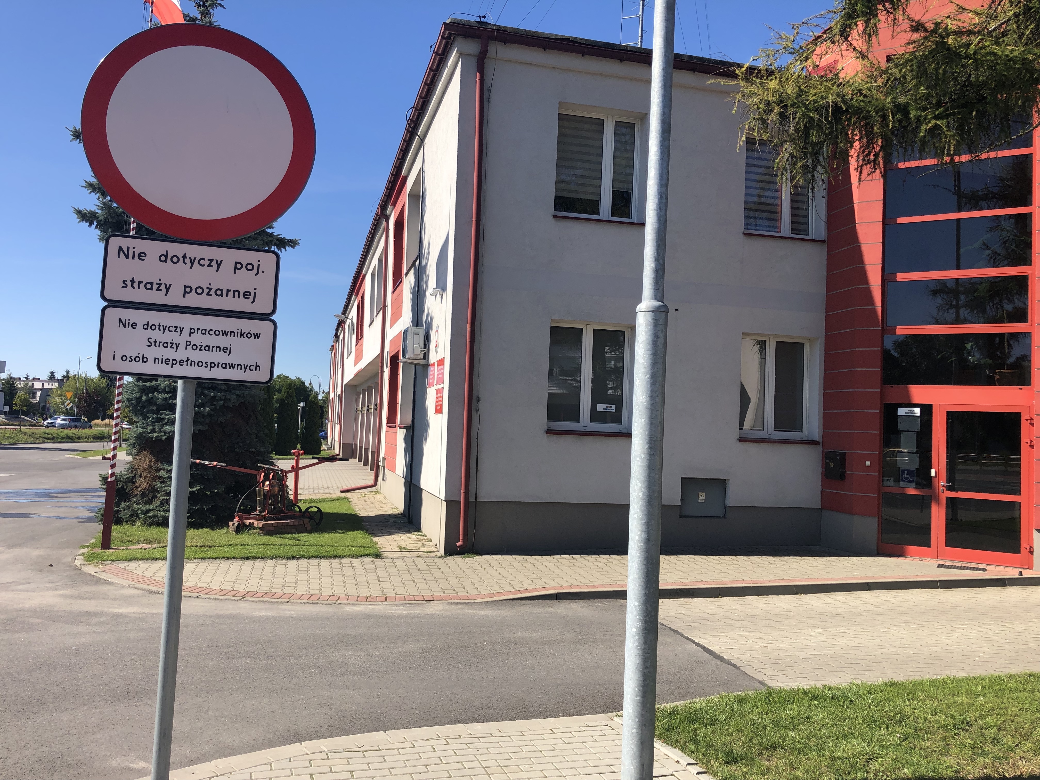 Na głównym planie znak zakaz wjazdu z tabliczkami: nie dotyczy pojazdów straży pożarnej oraz nie dotyczy pracowników Straży Pożarnej i osób niepełnosprawnych. W tle budynek komendy koloru szarego z czerwonymi elementami. Wejście do budynku.