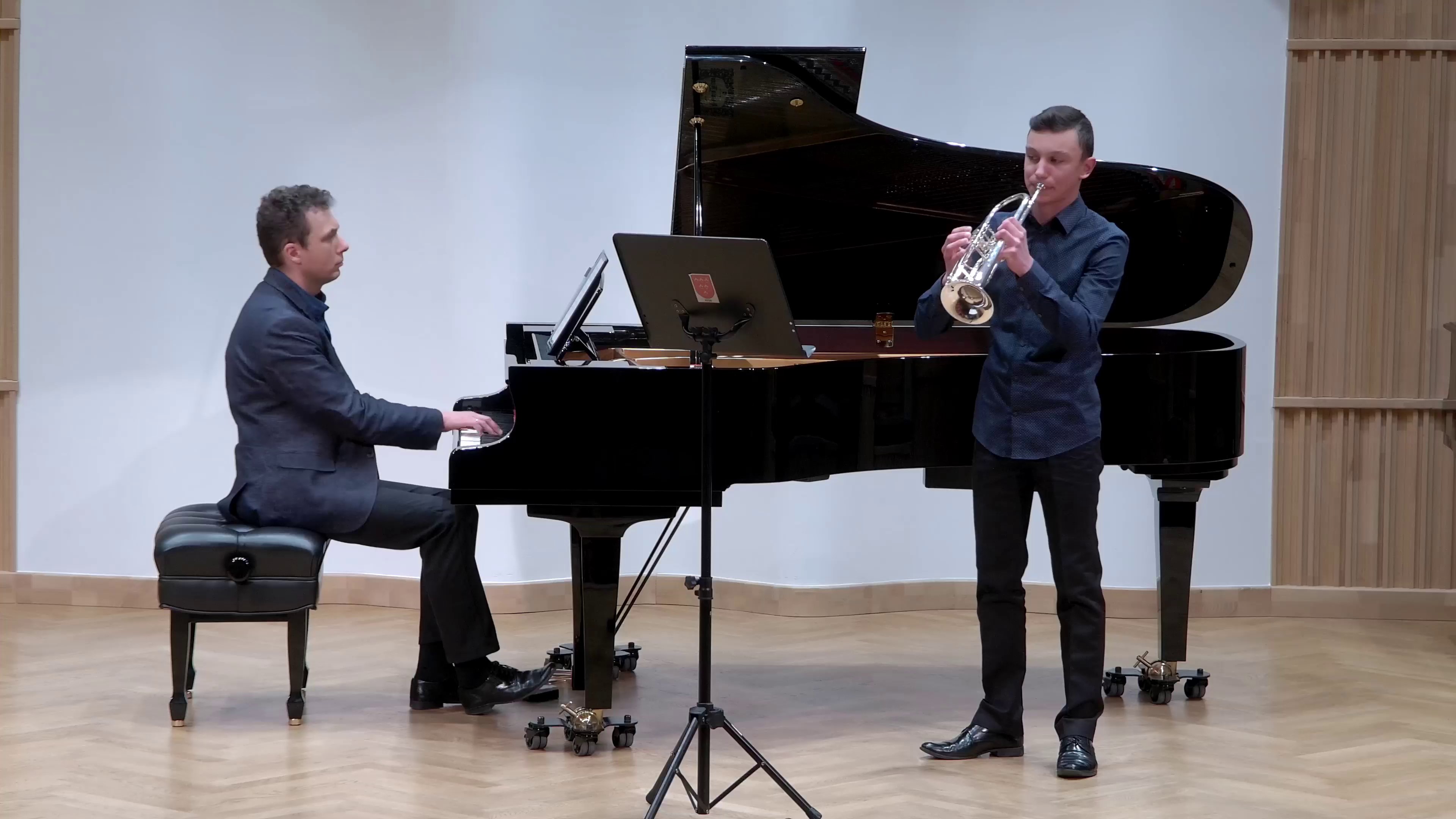 Zdjęcie. Na estradzie występuje młody trębacz z towarzyszeniem pianisty, który gra na dużym czarnym fortepianie.