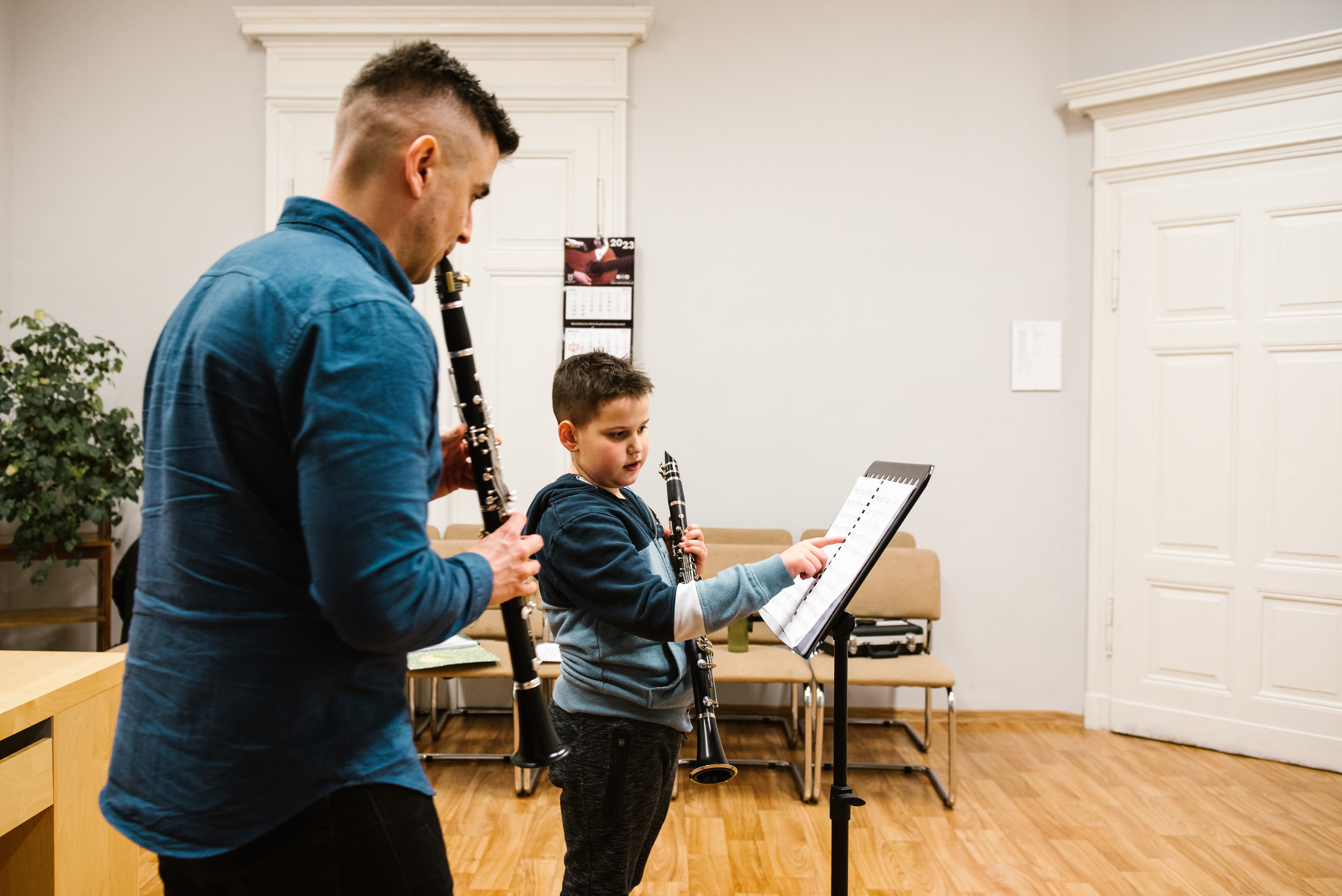 Na zdjęciu znajduje się nauczyciel Konrad Kołtun oraz uczeń Patryk Pawłowicz. Nauczyciel gra na klarnecie z nut znajdujących się na czarnym pulpicie. Uczeń trzyma w lewej ręce klarnet, prawą ręką wskazuje nuty palcem nauczycielowi. W tle stoją krzesełka klasowe, zielona roślina oraz biurko, na ścianie wisi kalendarz 