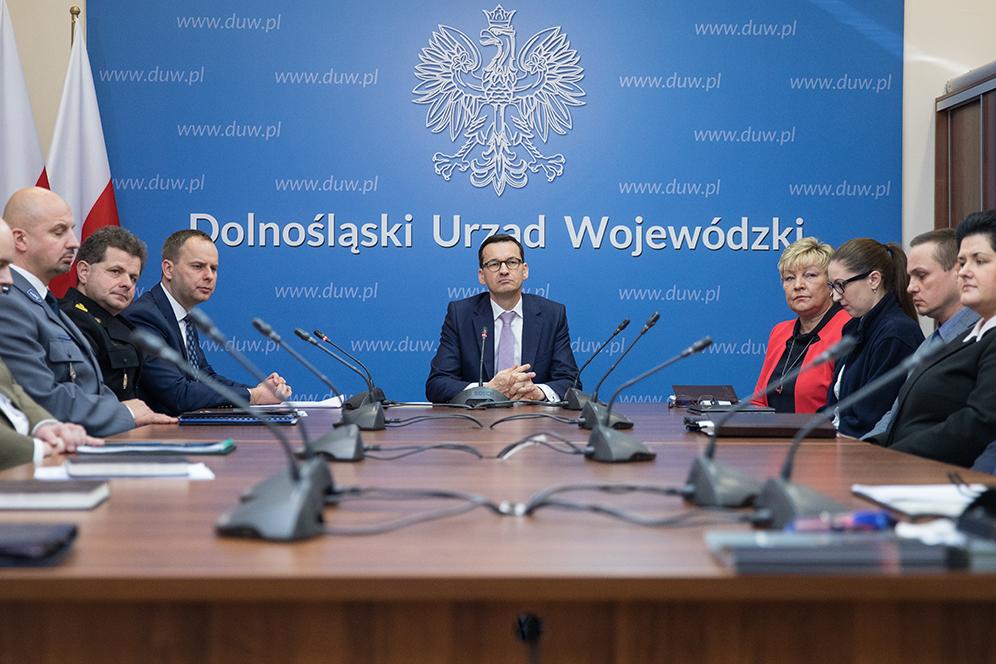 Premier Mateusz Morawiecki na tle niebieskiej ścianki z godłem.