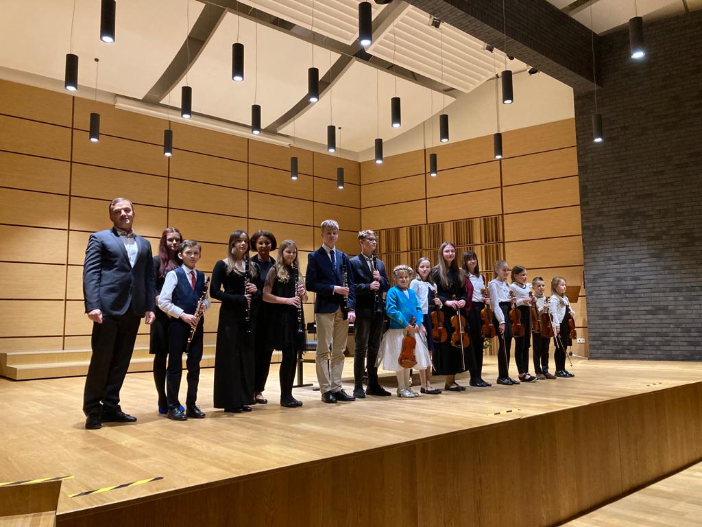 Uczniowie wraz z nauczycielami stojący w dwóch rzędach na scenie sali koncertowej. Z przodu uczniowie trzymają klarnety i skrzypce. Na górze widoczne wiszące lampy.