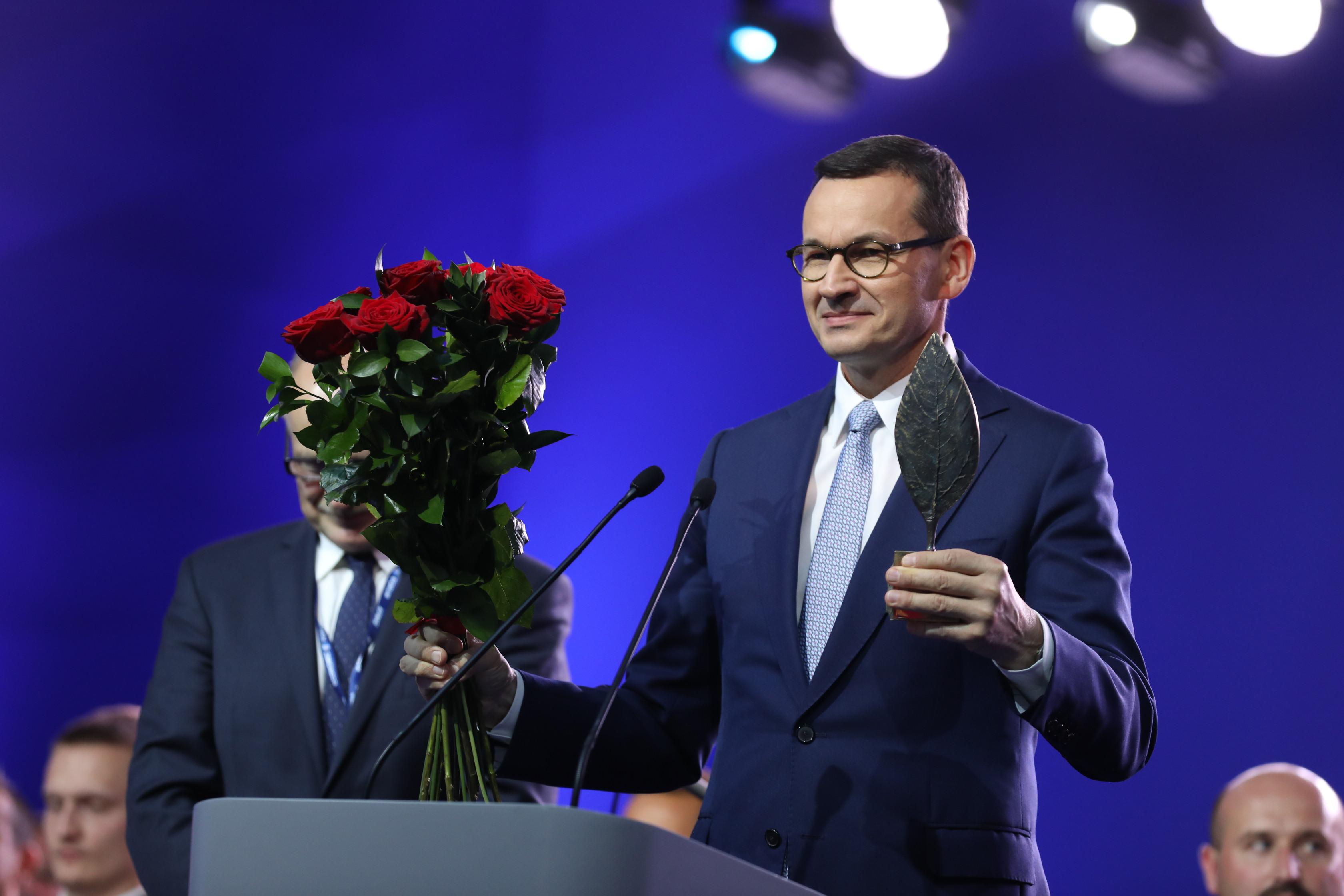 Premier Mateusz Morawiecki przemawia. W prawej ręce trzyma bukiet róż, w lewej statuetkę.