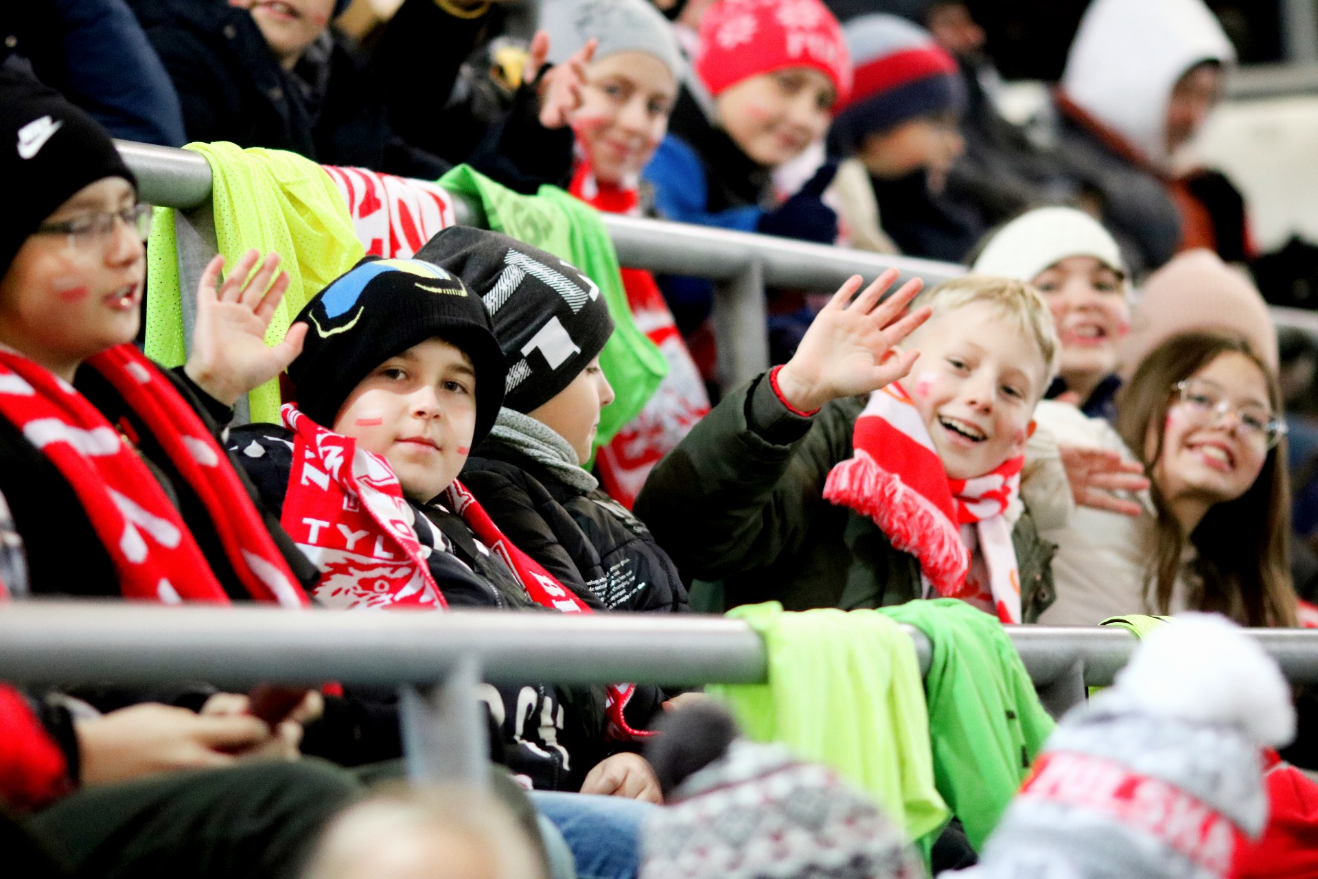 Kolorowo ubrane dzieci siedzą na trybunie stadionu, mają biało-czerwone szaliki, jeden chłopiec macha ręką.