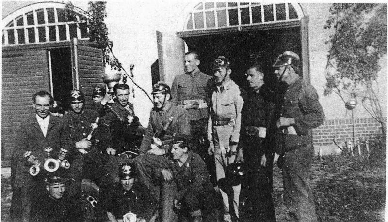 Czarno-białe zdjęcie przedstawia strażaków przed drzwiami strażnicy przy ulicy Piłsudskiego, część mężczyzn nosi hełmy, widać również rozdzielacz oraz prądownice proste,