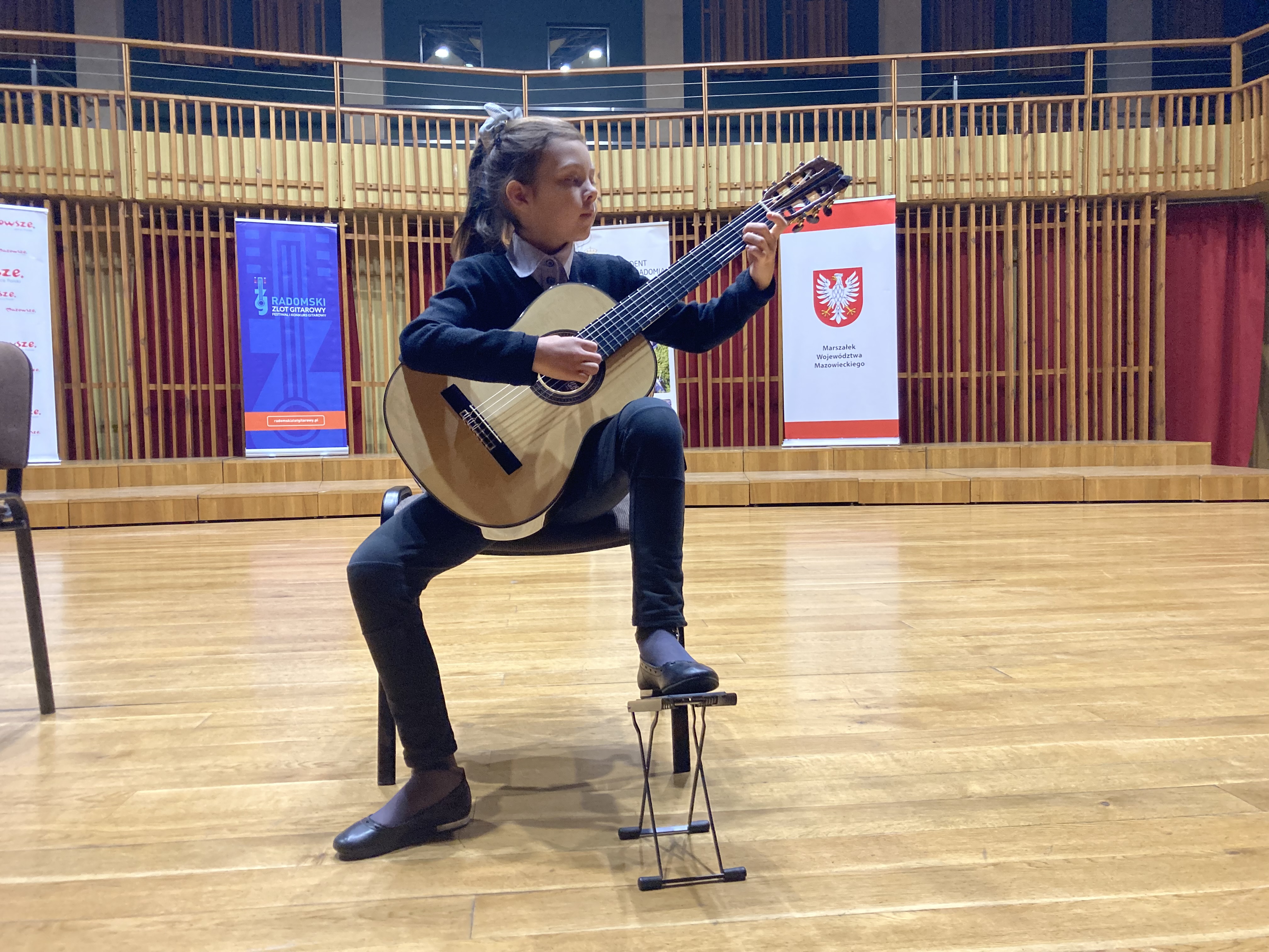 Zdjęcie przedstawia uczennice Lenę Torba grającą na scenie. W tle widać baner Radomskiego Zlotu Gitarowego oraz baner z napisem Marszałek Województwa Mazowieckiego nad którym widnieje godło Polski