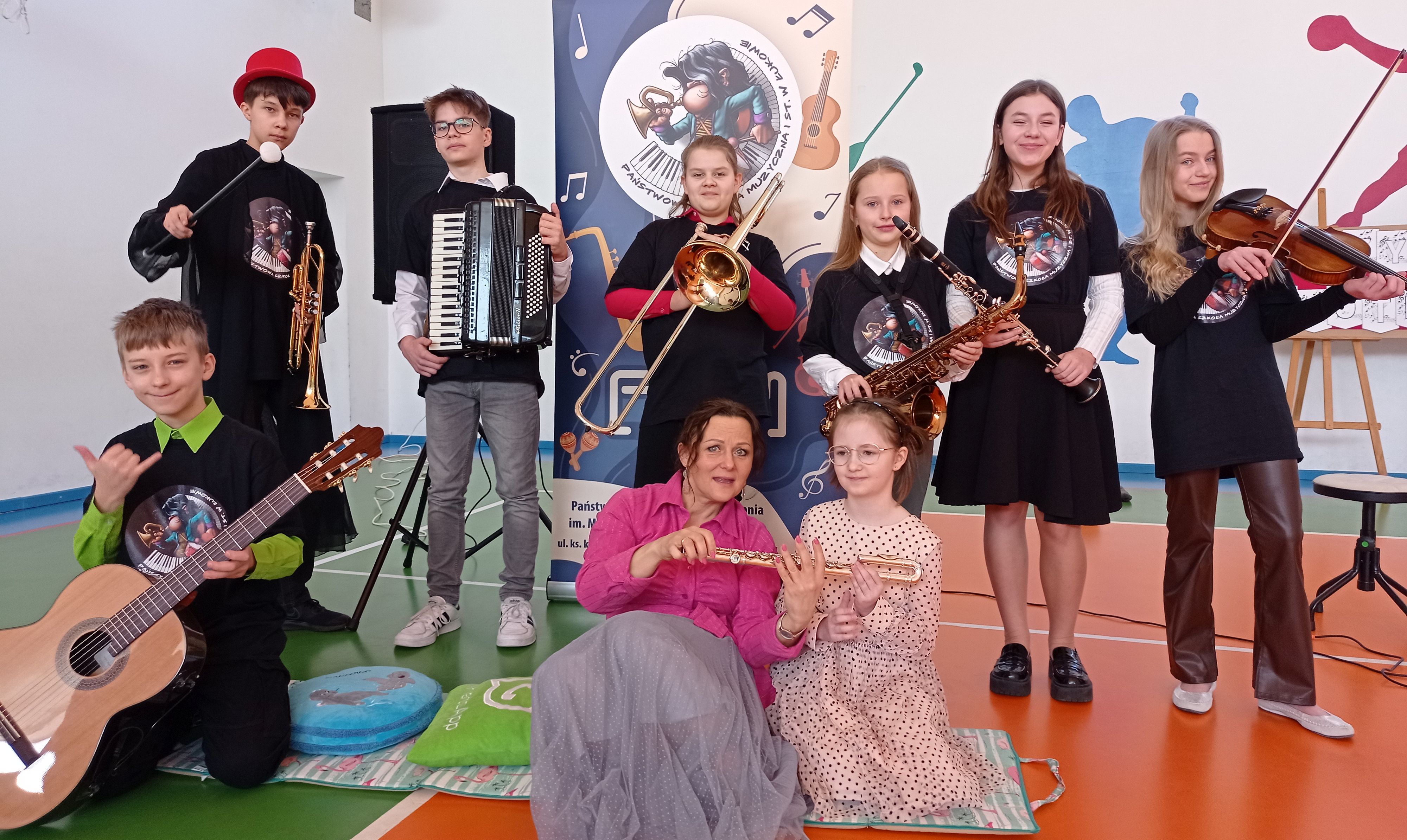 Ośmioro uczniów ze swoimi instrumentami i nauczycielka na tle banera reklamowego szkoły muzycznej w sali gimnastycznej Szkoły Podstawowej nr 4 w Łukowie