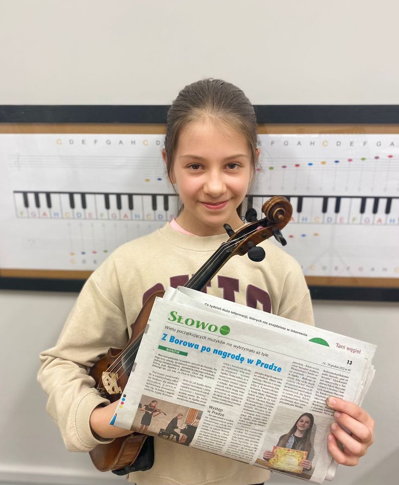 Zdjęcie przedstawia uczennicę Agatę Kostanowicz trzymającą skrzypce oraz gazetę, w której znajduje się artykuł o jej sukcesach. Dziewczynka ma na sobie beżową bluzę. Za nią widoczna jest tablica z klawiaturą fortepianu.