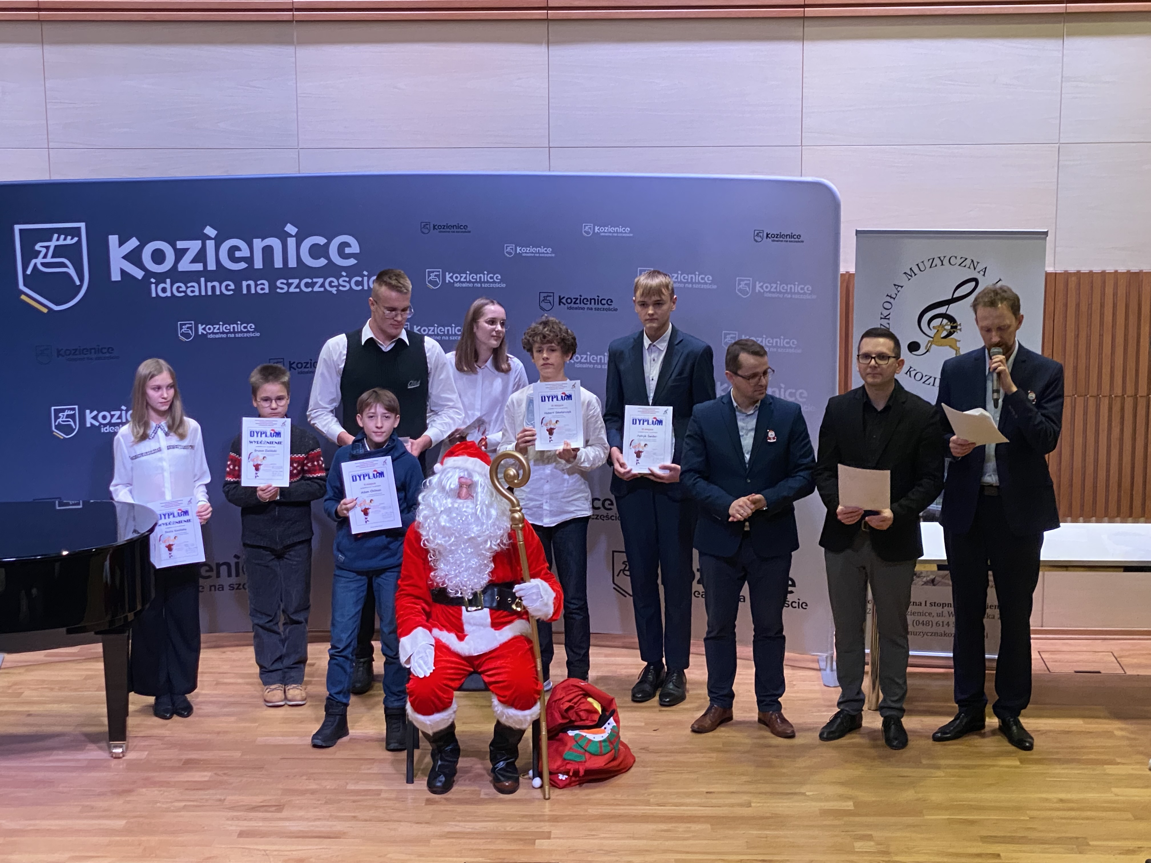 Zdjęcie przedstawia scenę, na której stoją laureaci konkursu trzymający w rękach dyplomy oraz jurorów. Przed nimi siedzi Mikołaj. W tle baner reklamowy w niebiskim kolorze z wieloma napisami "Kozienice" 