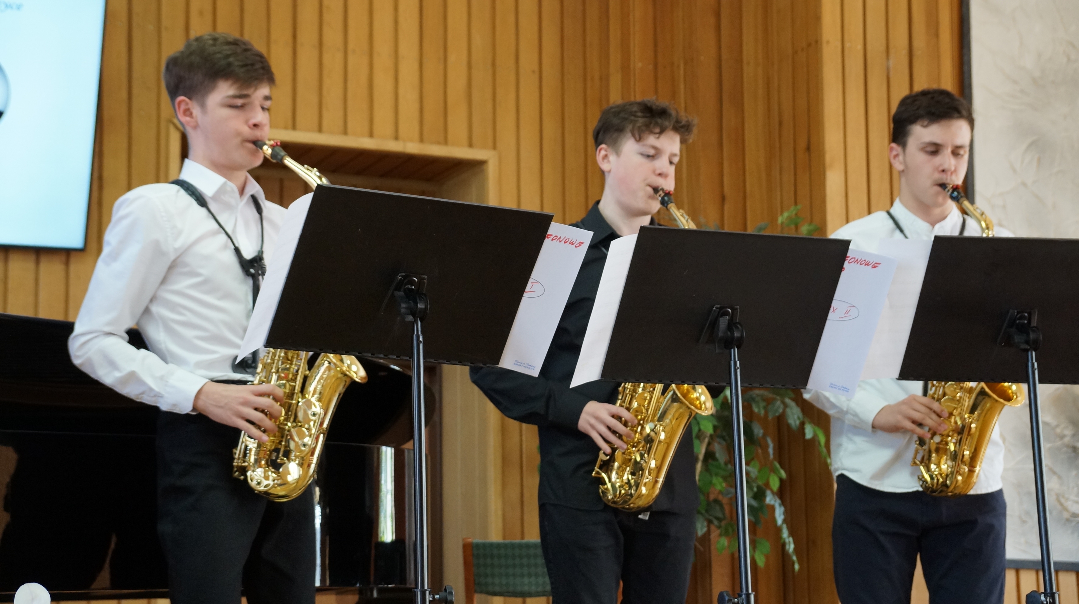 Troje uczniów ubranych na galowo grających na saksofonach w auli Państwowej szkoły Muzycznej