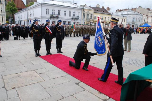 Na Sądeckim Rynku Komendant Miejski PSP w Nowym Sączu całuje sztandar przed jego przyjęciem od Komendanta Głownego PSP. W tle stoi poczet sztandarowy oraz pododdziały uczestniczące w uroczystości.