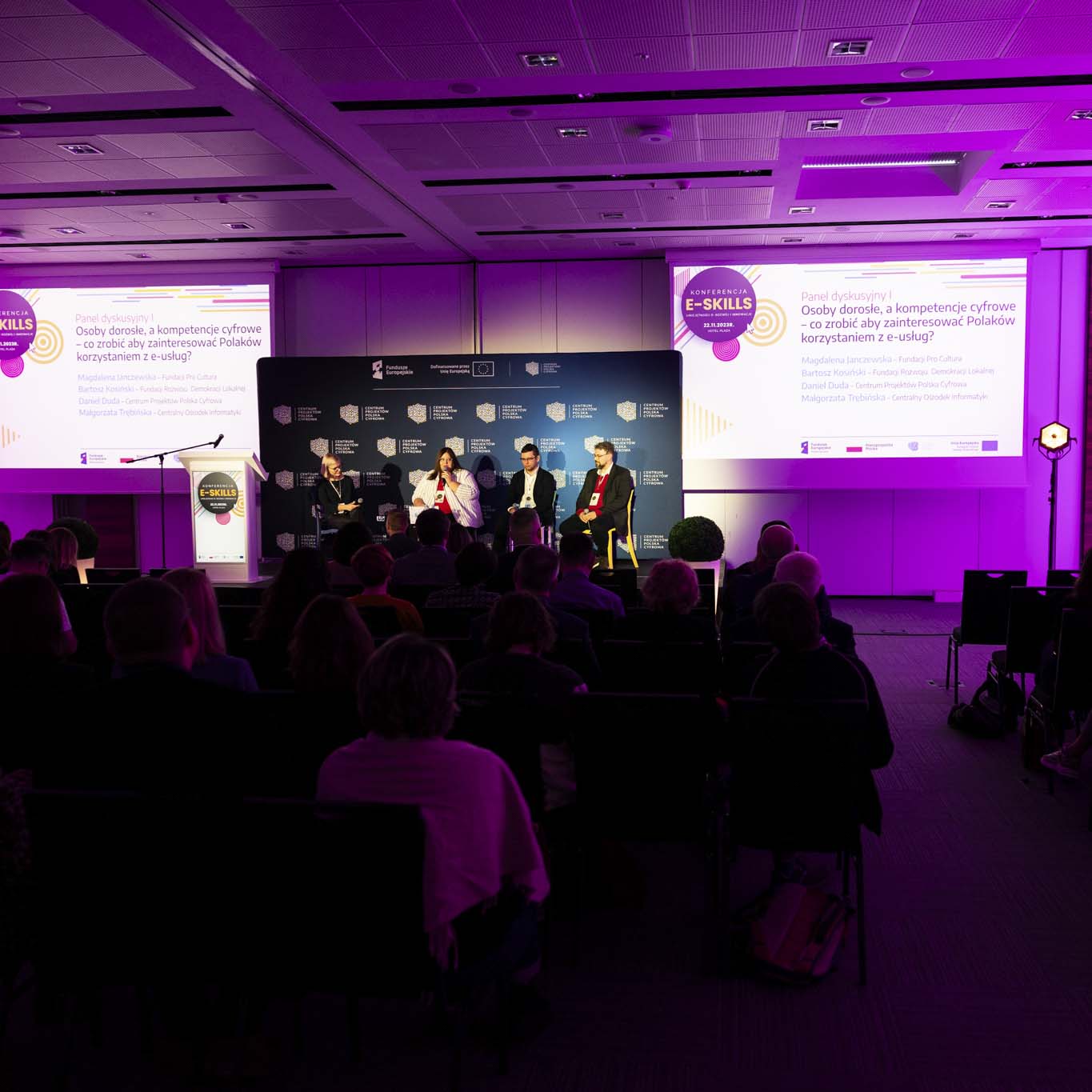 Czwórka panelistów siedzi na krzesłach na podium. Pani Magdalena Janczewska mówi do mikrofonu, reszta uczestników słucha wypowiedzi. Na pierwszy tle znajdują się uczestnicy konferencji.