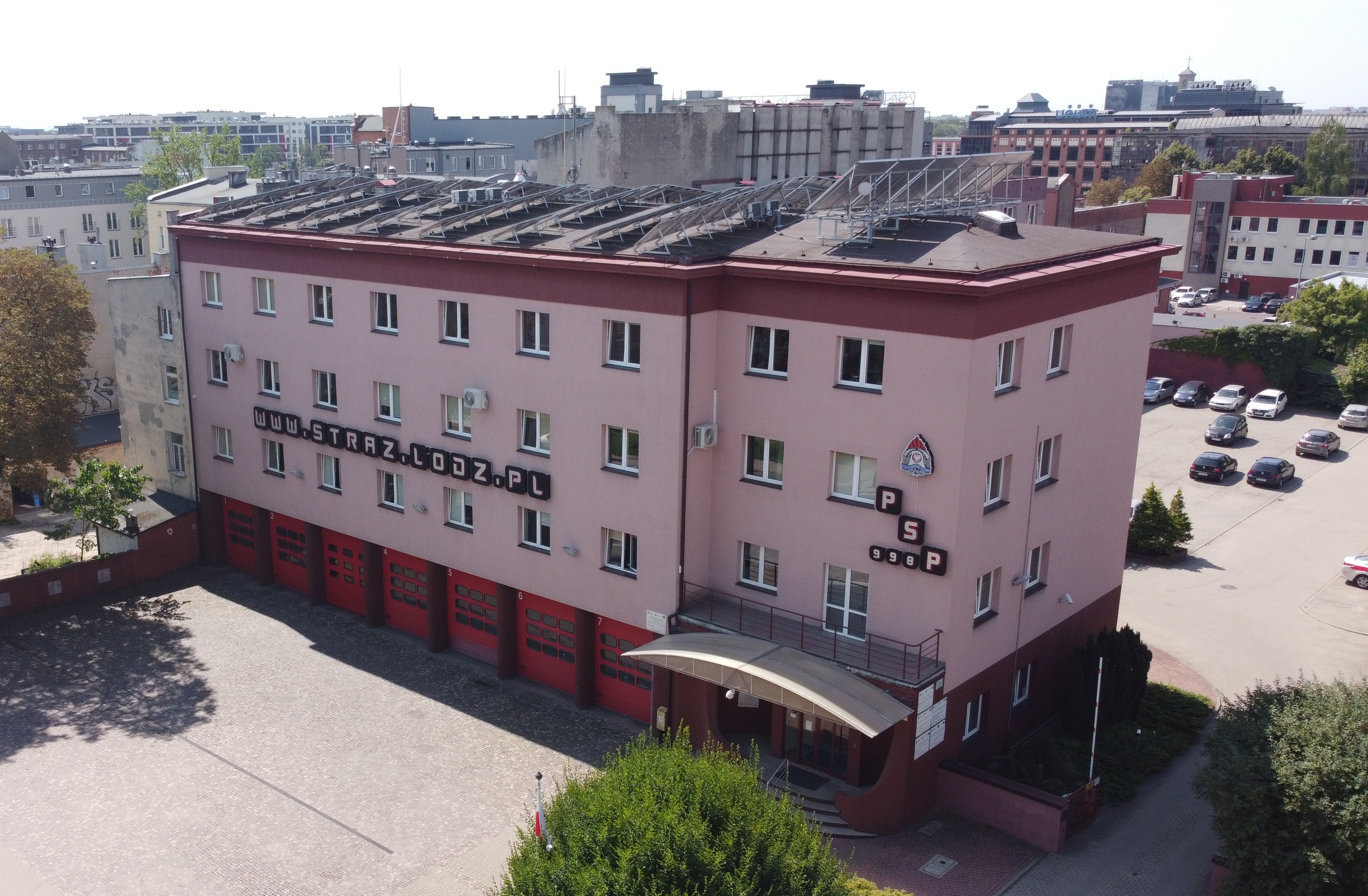 Zdjęcie przedstawia budynek Komendy Wojewódzkiej PSP w Łodzi, 3 piętrowy, elewacja w odcieniu różowego, na parterze 7 czerwonych bram garażowych, na dachu widać elementy instalacji fotowoltaicznej, pomiędzy okami na 1 i 2 piętrze napis www.straz.lodz.pl, nad wejściem na wysokości 2 piętra znajduje się logo Państwowej Straży Pożarnej, skrót PSP i nr telefonu 998