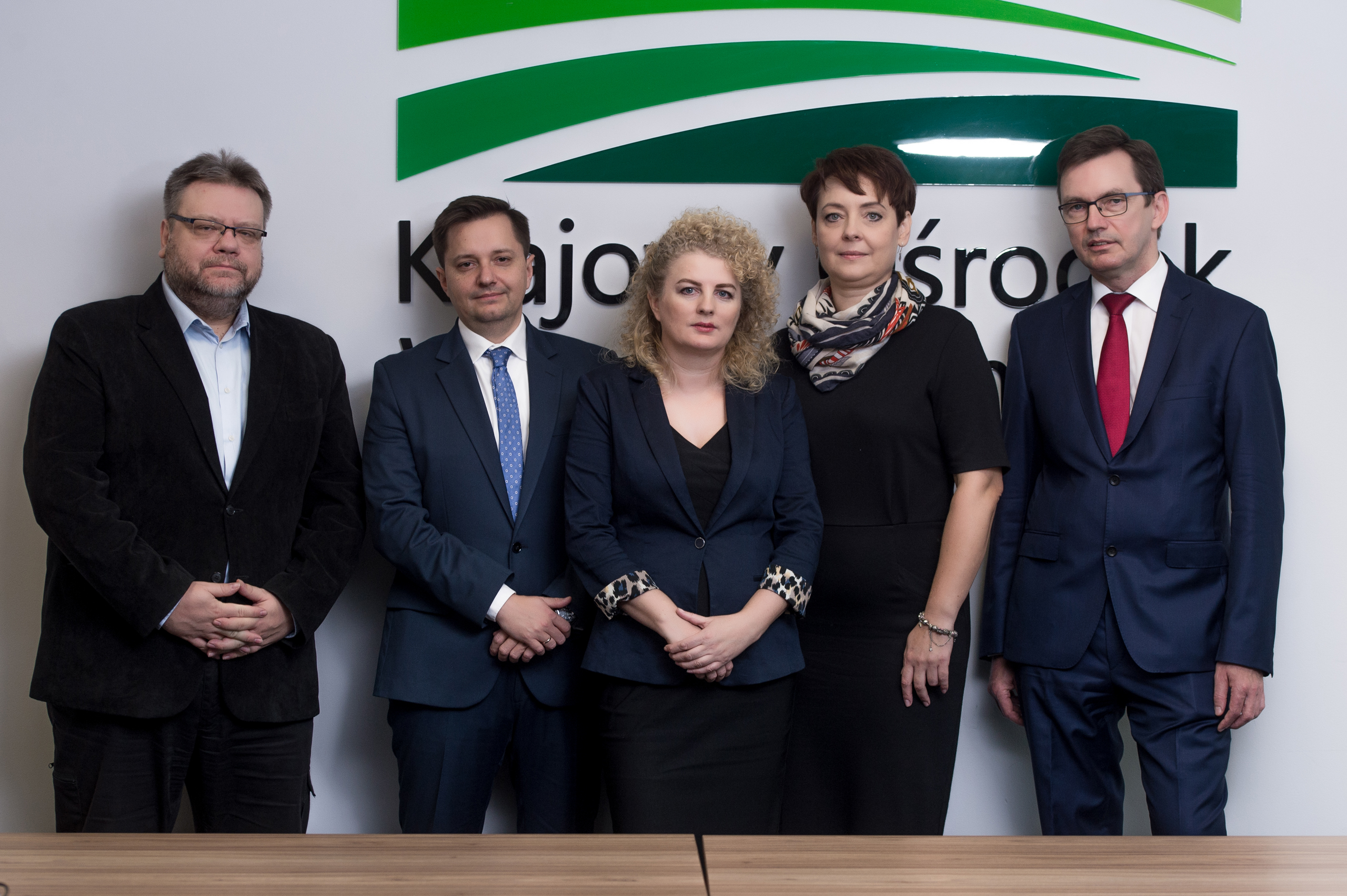 Na poziomym zdjęciu od lewej, w eleganckich ciemnych strojach, stoi 5 osób z zespołu projektowego, na tle częściowo zasłoniętego logotypu Krajowego Ośrodka Wsparcia Rolnictwa: Michał Milewski – p.o. kierownik Wydziału Projektów w Departamencie Innowacji KOWR, Mateusz Balcerowicz – dyrektor Departamentu Innowacji (kierownik projektu wobec NCBR), Bożena Tybora – główny specjalista w Wydziale Projektów, Departament Innowacji, Elżbieta Jagłowska-Zawadka – główny specjalista w Wydziale Projektów, Departament Innowacji, Wojciech Kędzia – zastępca Dyrektora Generalnego KOWR.