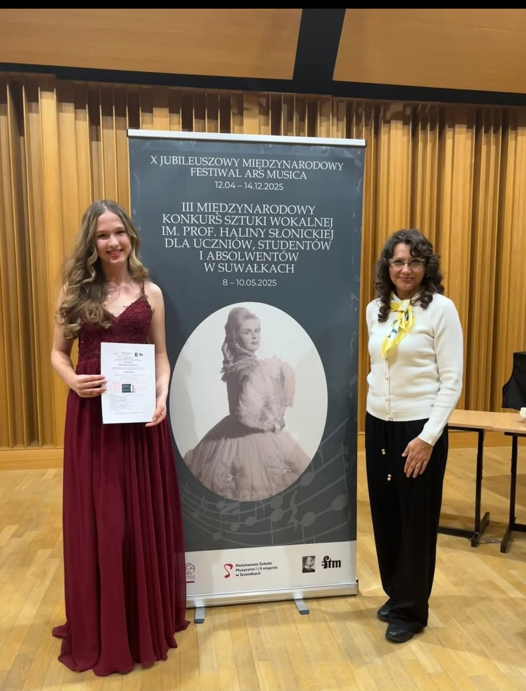 Amelia Szmidt i Monika Brewczak - III Międzynarodowy Konkurs Sztuki Wokalnej w Suwałkach. Uczennica i nauczyciel stojące przy banerze reklamowym konkursu na scenie sali koncertowej.