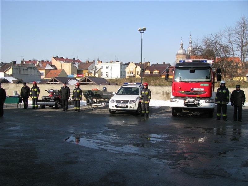 Na placu stoją przekazywane dwa samochody jeden lekki koloru białego a drugi średni koloru czerwonego.Obok stoją przyjmujący je strażacy. W tle widać budynki z okolic Sądeckiego Rynku oraz wieże Bazyliki świętej Małgorzaty.