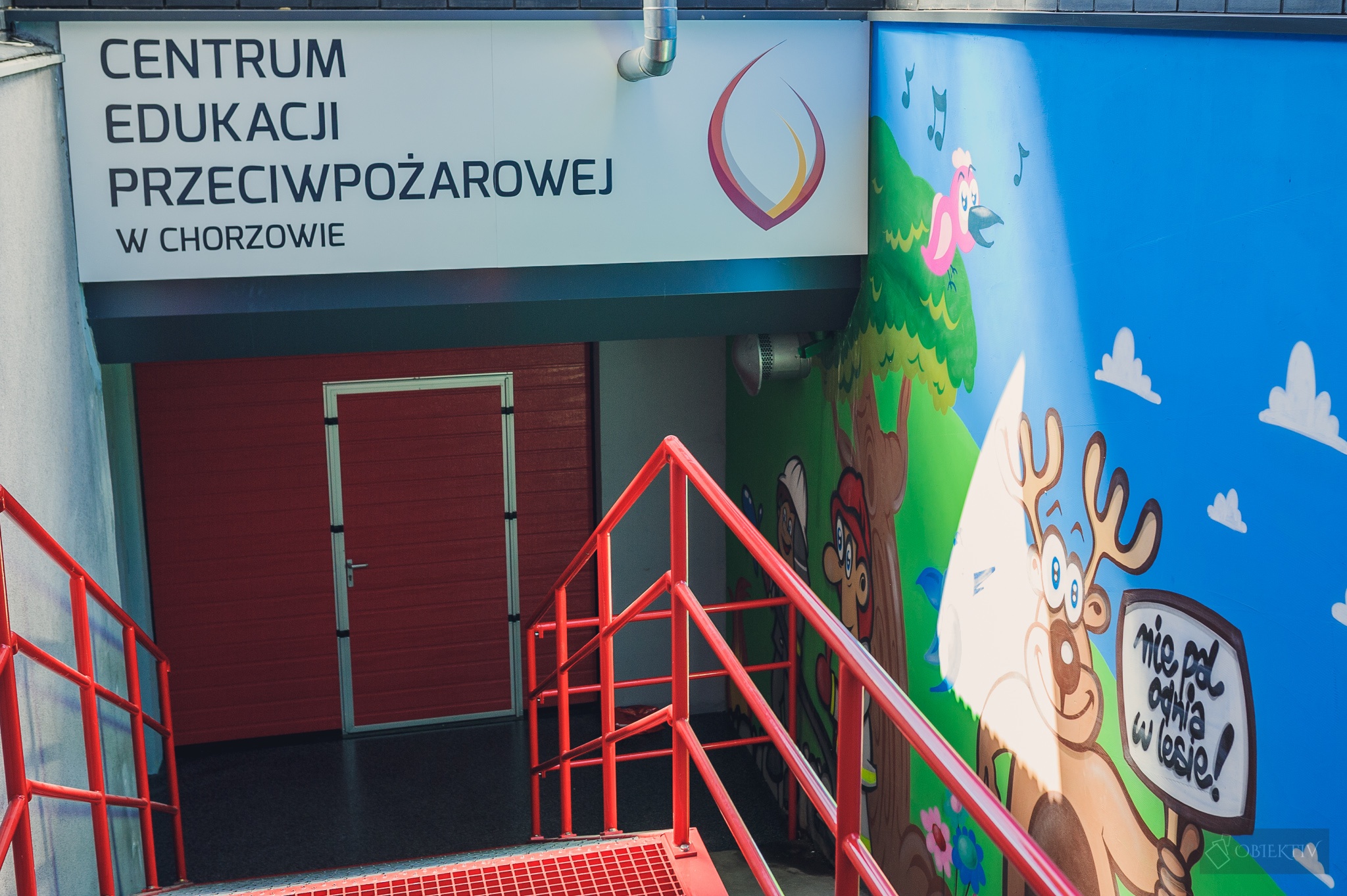 Zdjęcie przedstawia wejście do Centrum Edukacji Pożarowej. Czerwona brama wejściowa nad którą jest napis centrum edukacji pożarowej w Chorzowie
