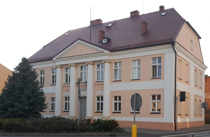 Zdjęcie przedstawia budynek PSM. Widoczne są dwie ściany budynku, front oraz ściana boczna. Na froncie znajdują się cztery kolumny rozdzielona oknami a na parterze między drugą a trzecią kolumną znajdują się drzwi główne do budynku. Przed budynkiem z lewej strony zdjęcia znajduje się wysokie drzewo iglaste a w dole na środku klomb zieleni.