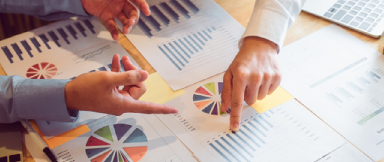 Na obrazku widać dwie osoby siedzące przy stole. Patrzą na kartki z wykresami. Jedna osoba pokazuje palcem na wykres. Druga osoba pokazuje coś ręką. Na stole leży też laptop.