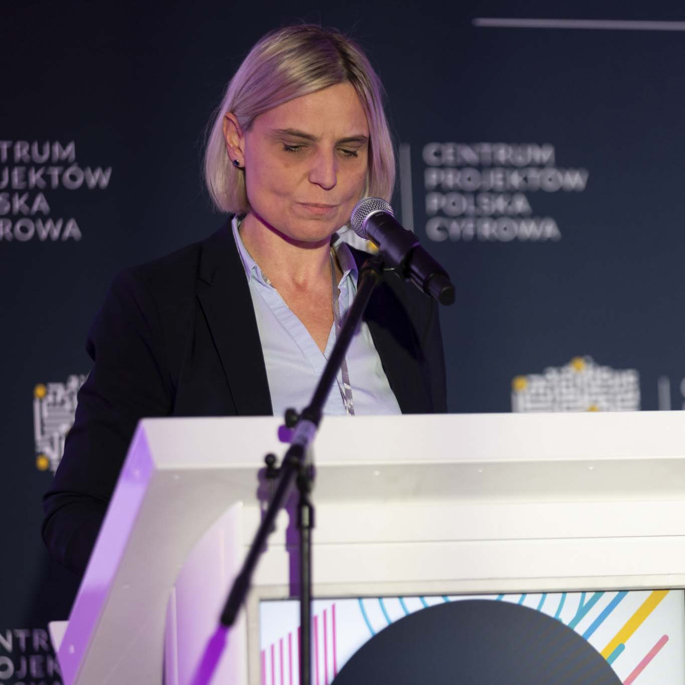 Przy mównicy stoi Pani Monika Eilmes. Wzrok kieruje w dół. Mówi do mikrofonu.