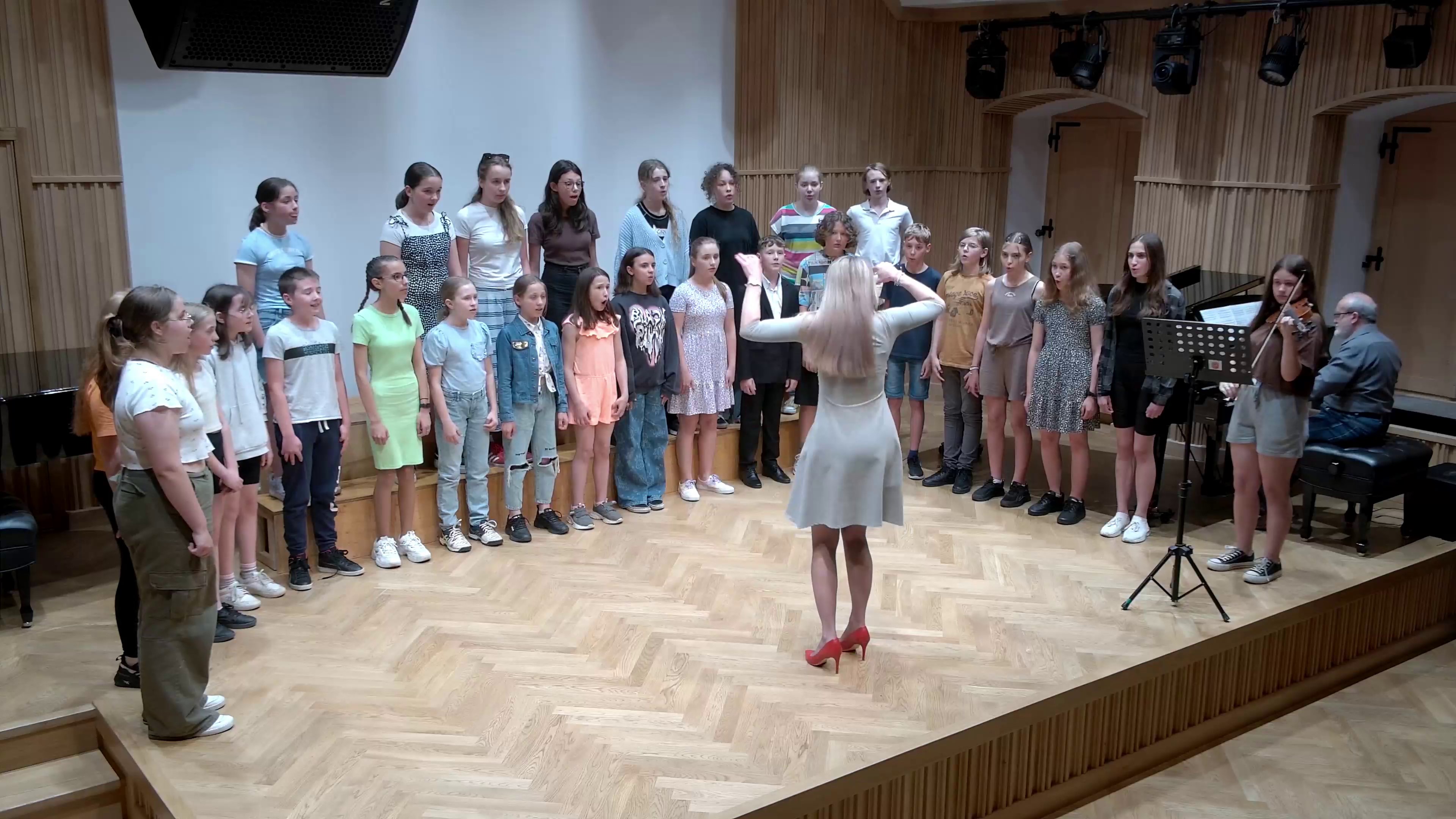 Zdjęcie. Na estradzie sali koncertowej występuje chór dziecięcy.. Z prawej strony widać również pianistę akompaniującego chórowi. Zespołem dyryguje pani dyrygent.