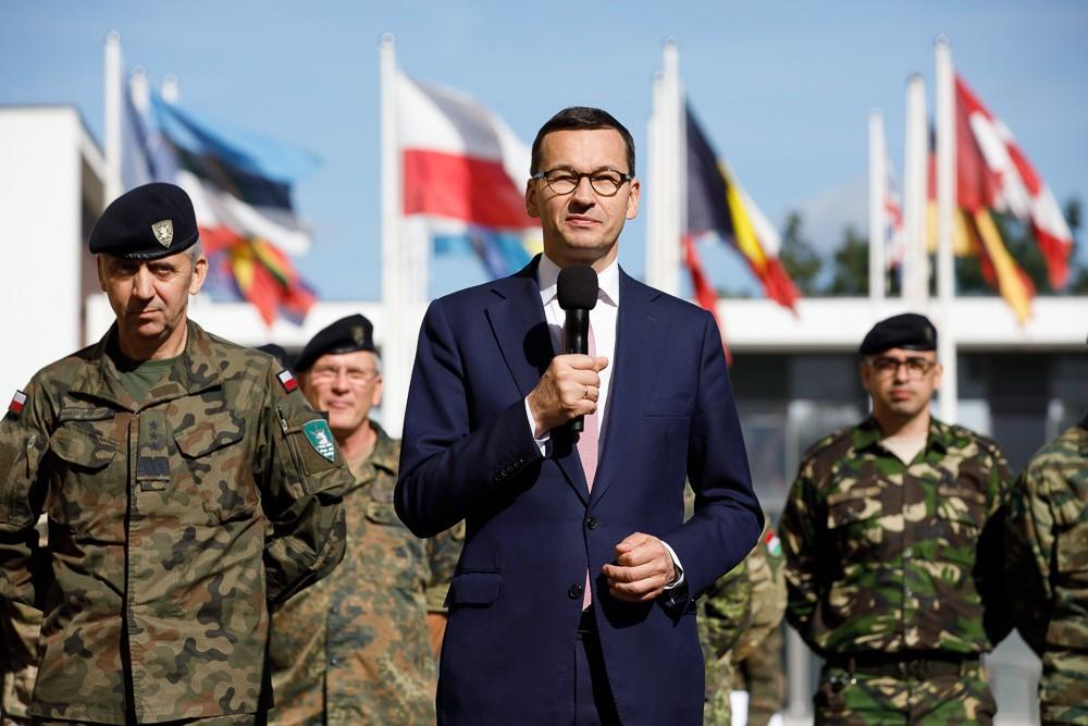Premier Mateusz Morawiecki z żołnierzami, a w tle flagi.