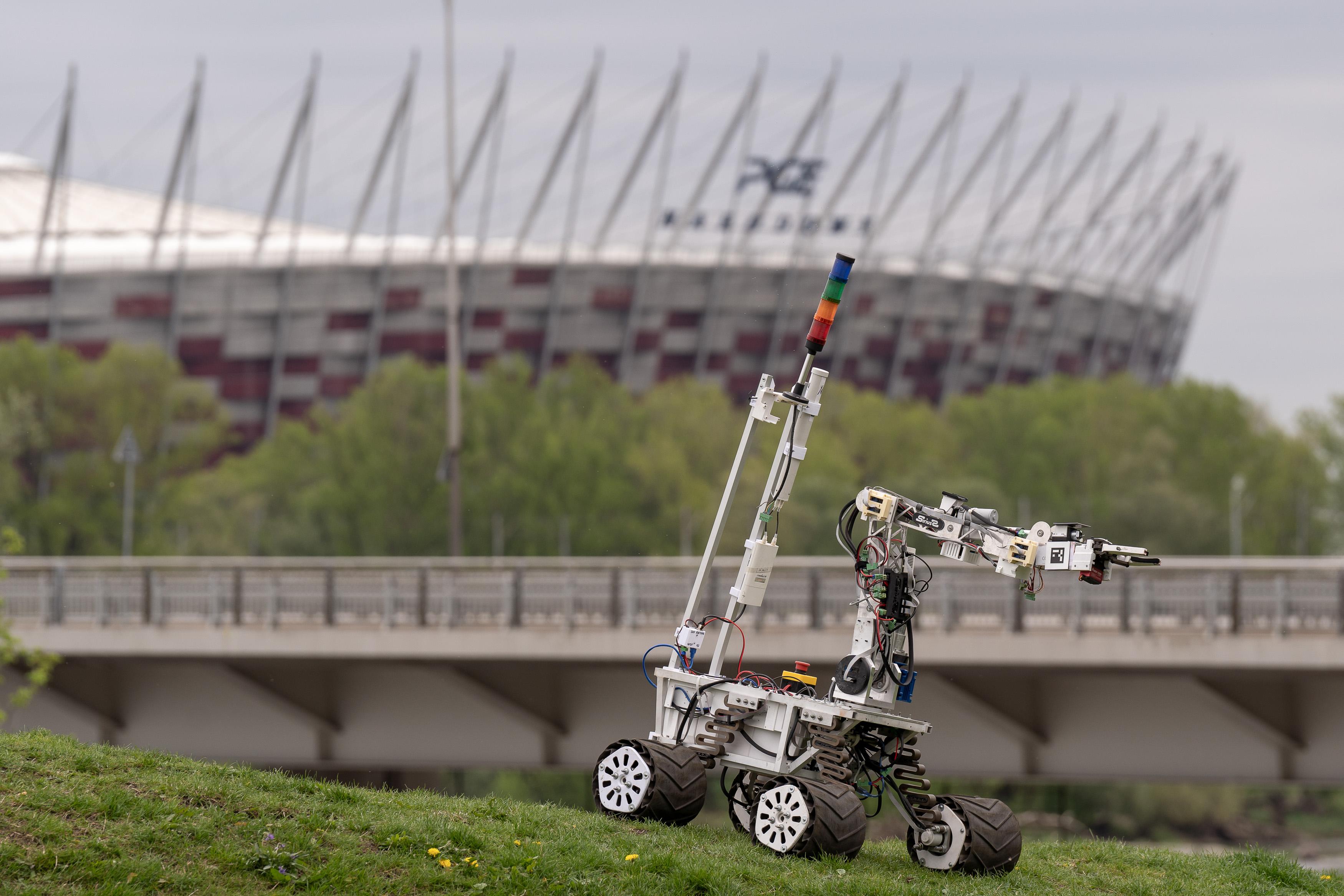 Łazik stoi na wzniesieniu na trawie, w tle most i Stadion Narodowy