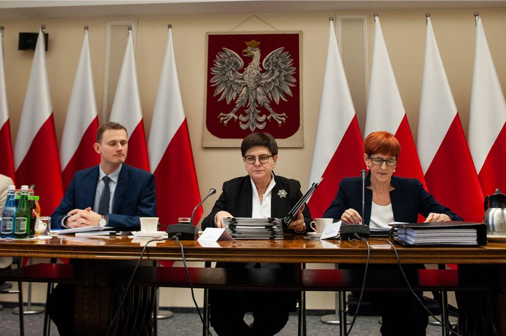 Wicepremier Beata Szydło i minister rodziny, pracy i polityki społecznej Elżbieta Rafalska podczas posiedzenia komisji, a w tle godło i biało-czerwone flagi.
