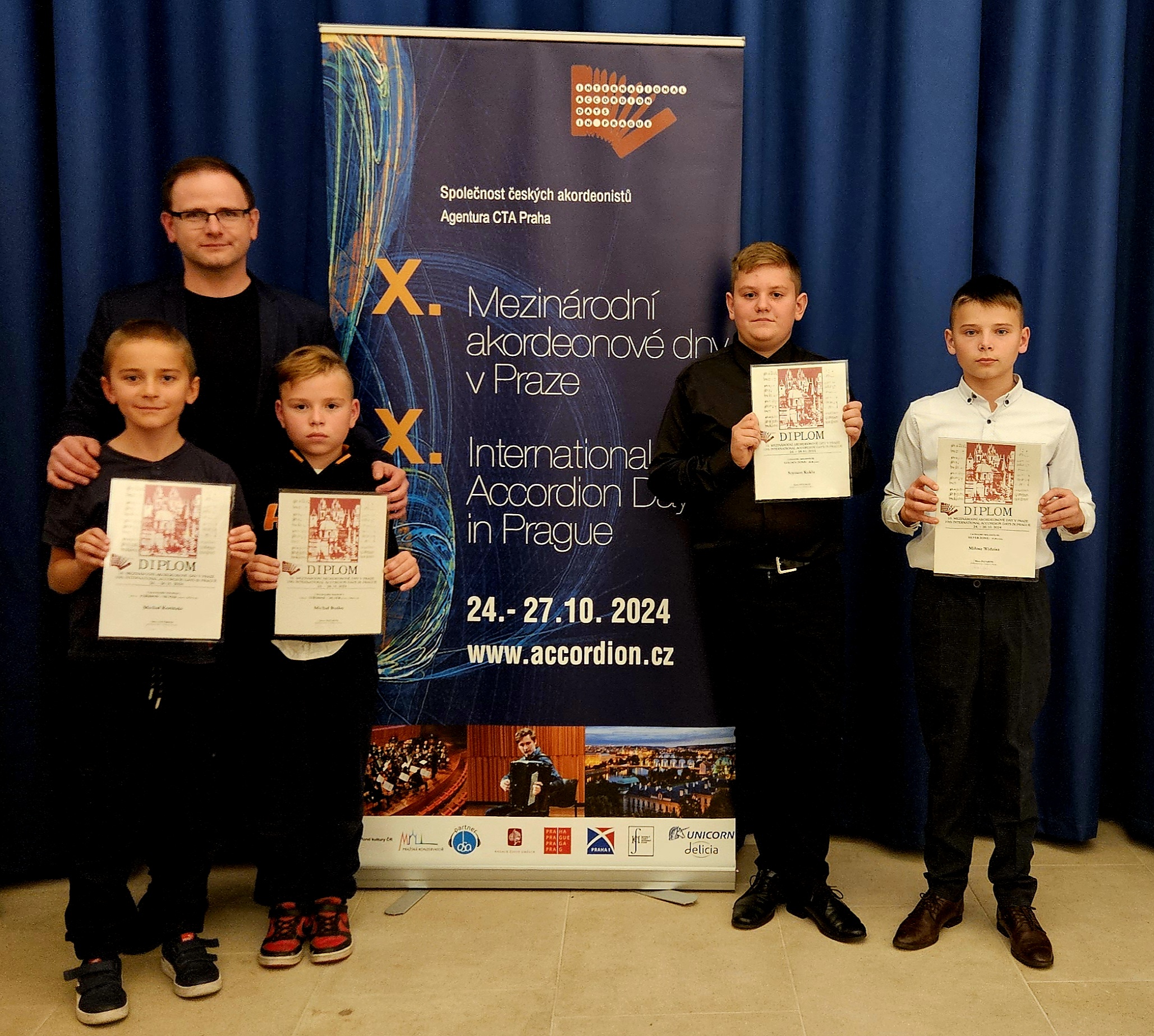 Stojący obok rollup-a uczniowie prezentujący zdobyte dyplomy wraz z nauczycielem. Za ich plecami granatowa kotara