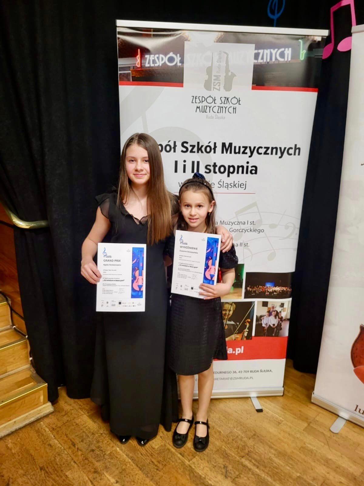 Na zdjęciu znajdują się dwie uśmiechnięte uczennice w czarnych sukienkach, w rękach trzymają dyplomy konkursowe. Za nimi znajduje się szkolny baner, w tle widnieje czarna kotara. 