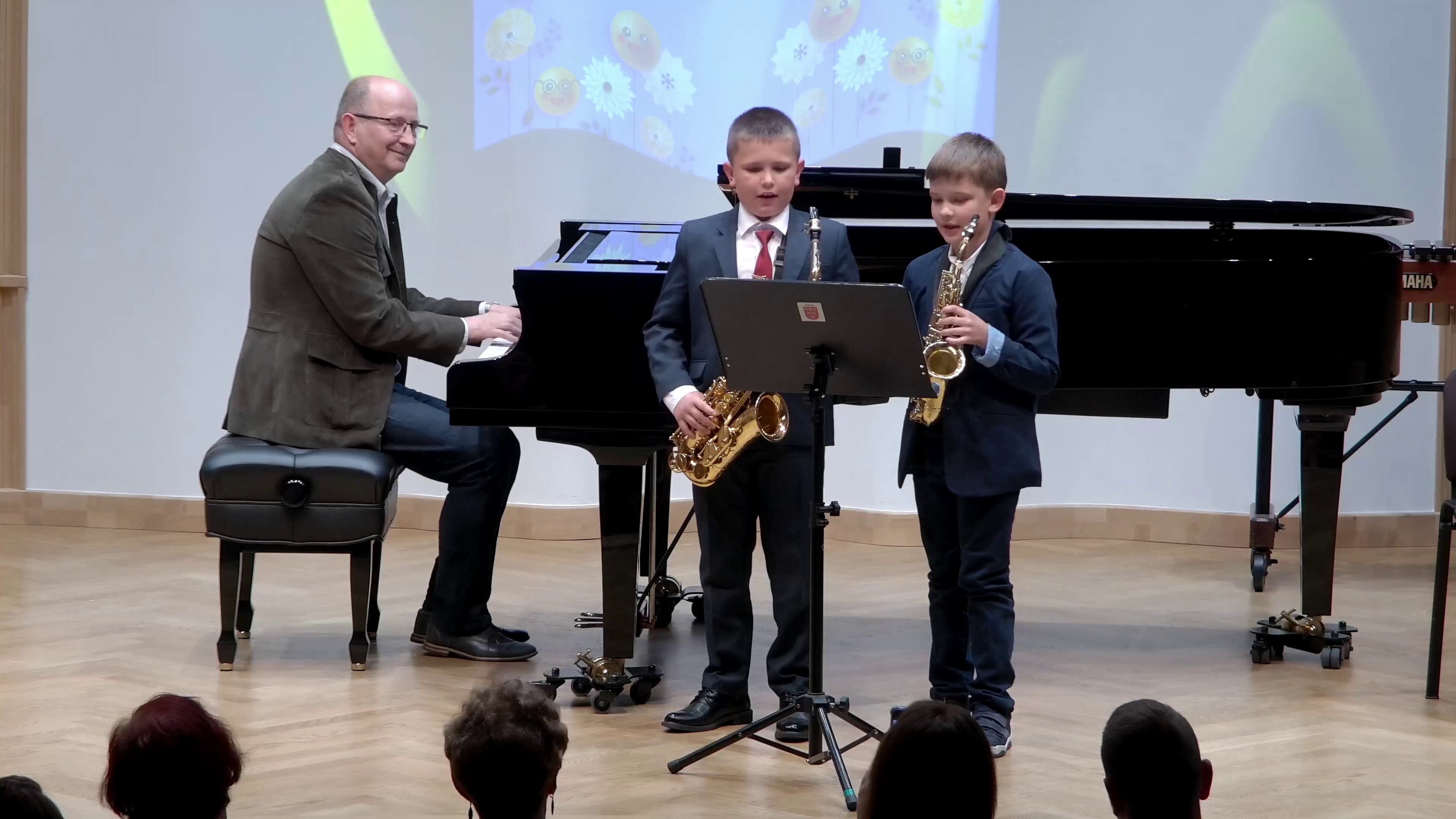 Zdjęcie. na estradzie występuje duet młodych saksofonistów ze swoim nauczycielem, który towarzyszy im grając na fortepianie. Występowi wsłuchuje się publiczność.