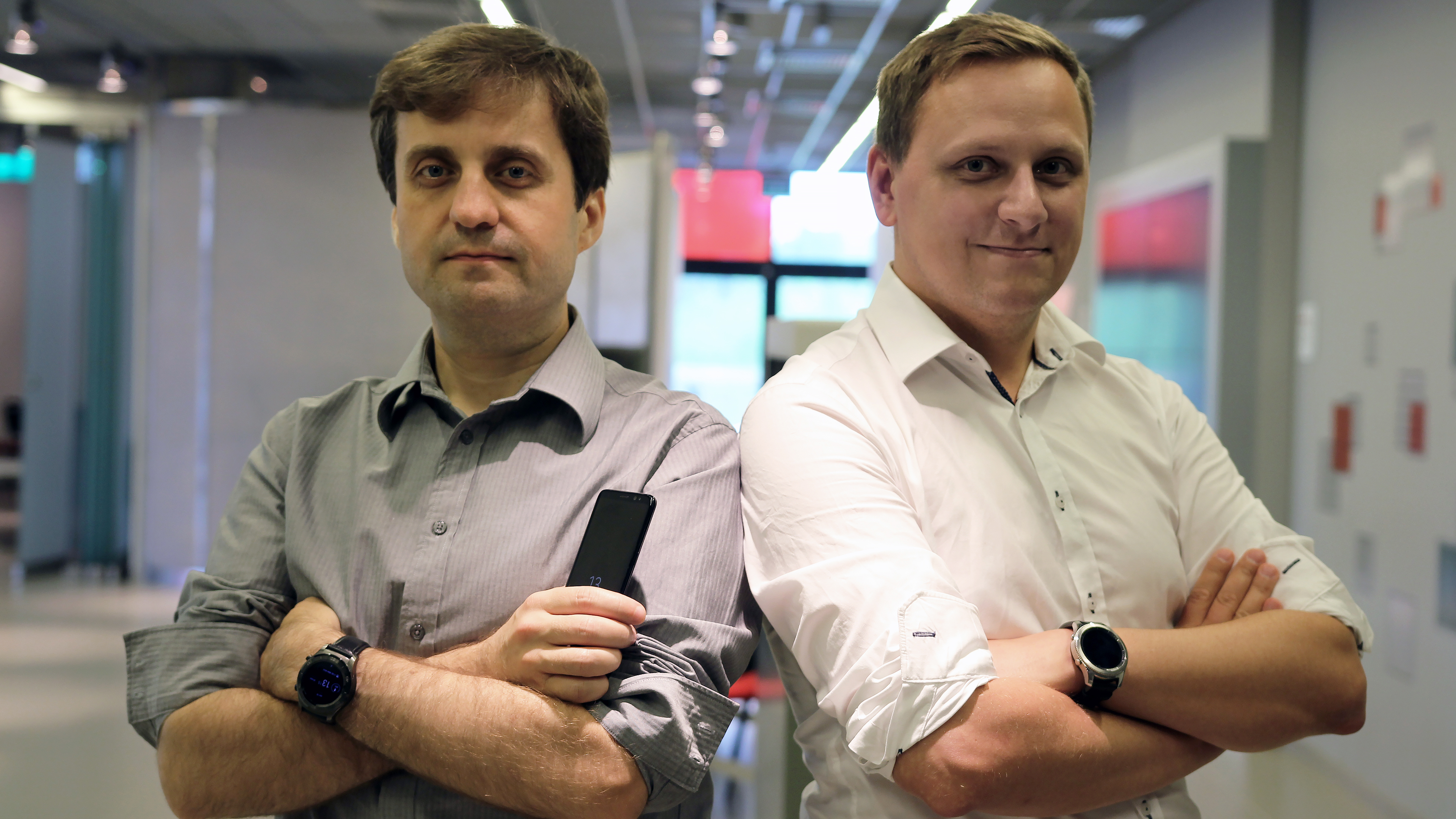 W ciasnym kadrze od pasa w górę dwóch mężczyzn w wieku około 30 lat, w jasnych koszulach, na tle dużej jasnej sali. Każdy z mężczyzn z rękami założonymi jedna na drugą. Mężczyzna z prawej strony trzyma w prawej dłoni czarny telefon komórkowy.