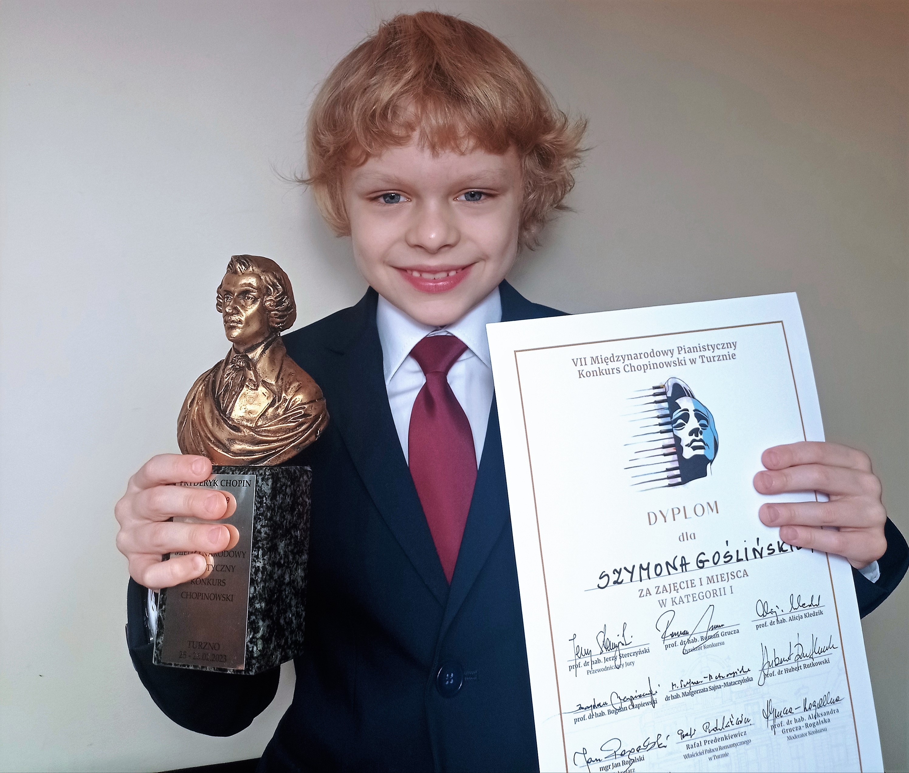 zdjęcie od pasa w górę Szymona Goślińskiego trzymającego dyplom w jednej ręce oraz statuetkę z popiersiem Chopina w drugiej, całość na białym tle