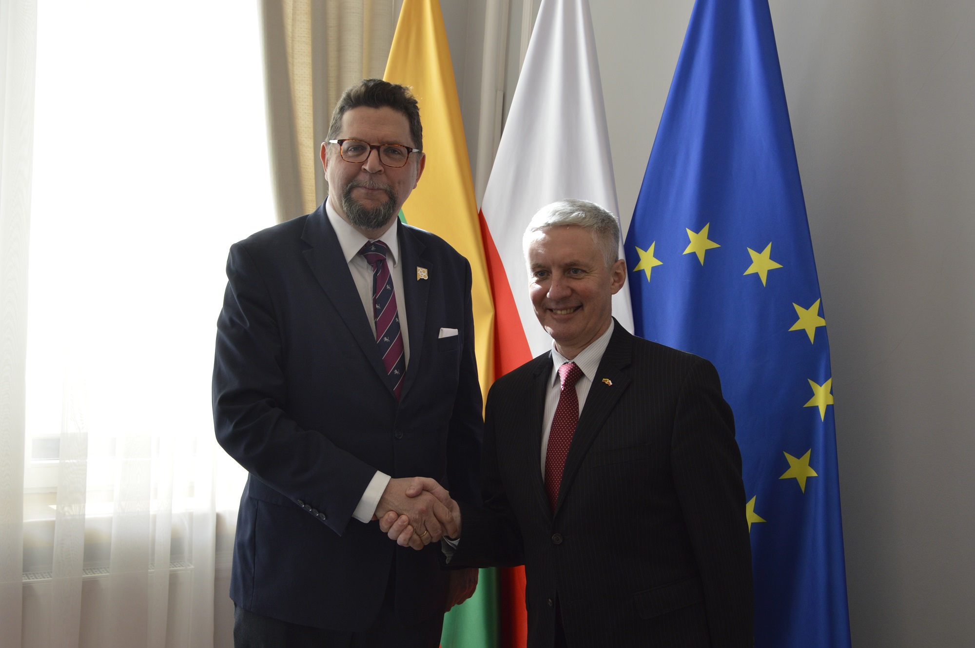 Wiceminister Szeptycki stoi obok mężczyzny w garniturze, podaje mu rękę, za nimi pod białą ścianą flagi Polski, Litwy i UE, z lewej zasłonięte firanką okno.
