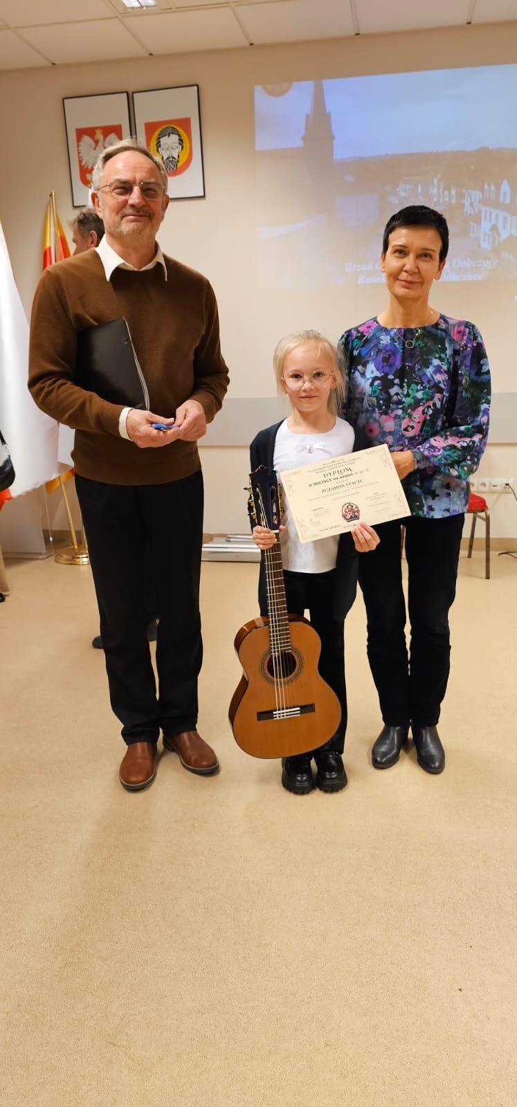 Zuzanna Stach z jurorami VII Ogólnopolskiego Konkursu Gitarowego „Młodzi czarodzieje gitary” w Dobczycach. Trzy osoby na zdjęciu w sali przesłuchań konkursowych.