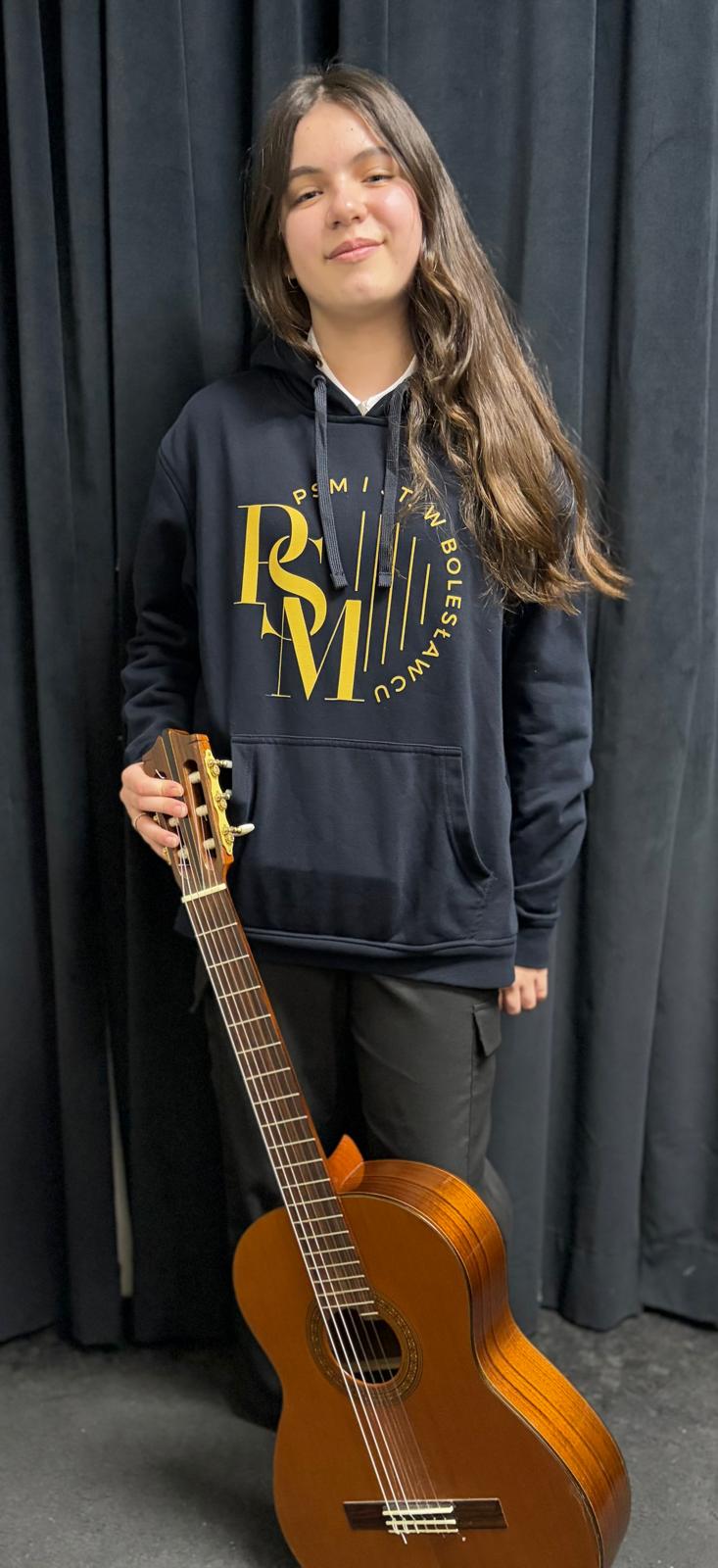 Zdjęcie pozowane, przedstawiające uczennicę ubraną w granatową bluzę z logo szkoły oraz trzymającą w rękach, opartą o nogi - gitarę klasyczną.