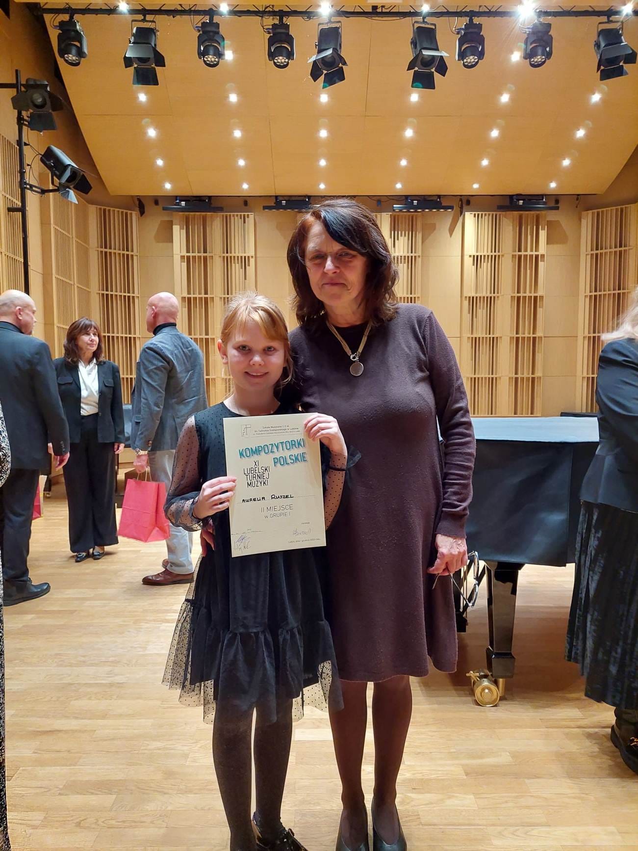 Na zdjęciu uczennica Aurelia Ruszel stoi w sali koncertowej trzymając dyplom za zajęcie II miejsca. Obok stoi pani Iwona Siedlaczek