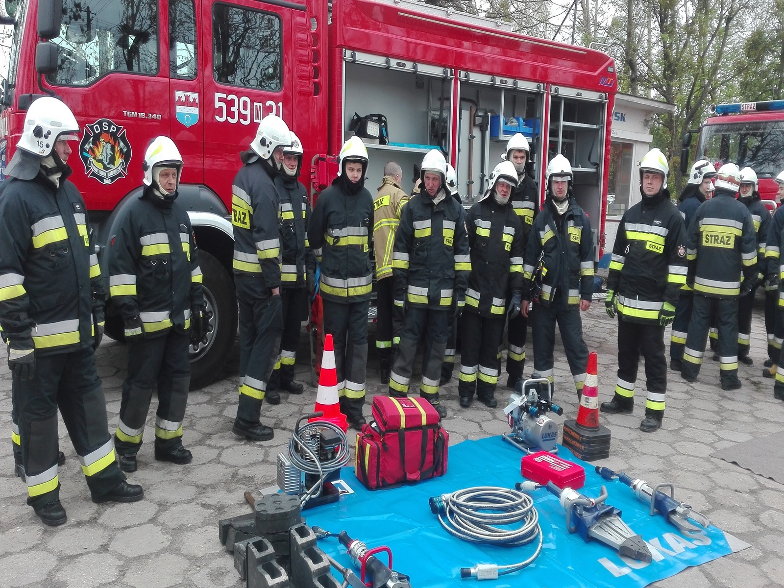 Zdjęcie przedstawia grupę strażaków OSP na tle samochodów pożarniczych, zebranych wokół sprzętu ratowniczego leżącego na niebieskiej plandece.