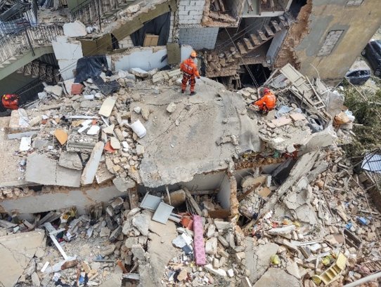 Zdjęcie przedstawia trzech ratowników szukających w zniszczonym wybuchem budynku ofiar i poszkodowanych