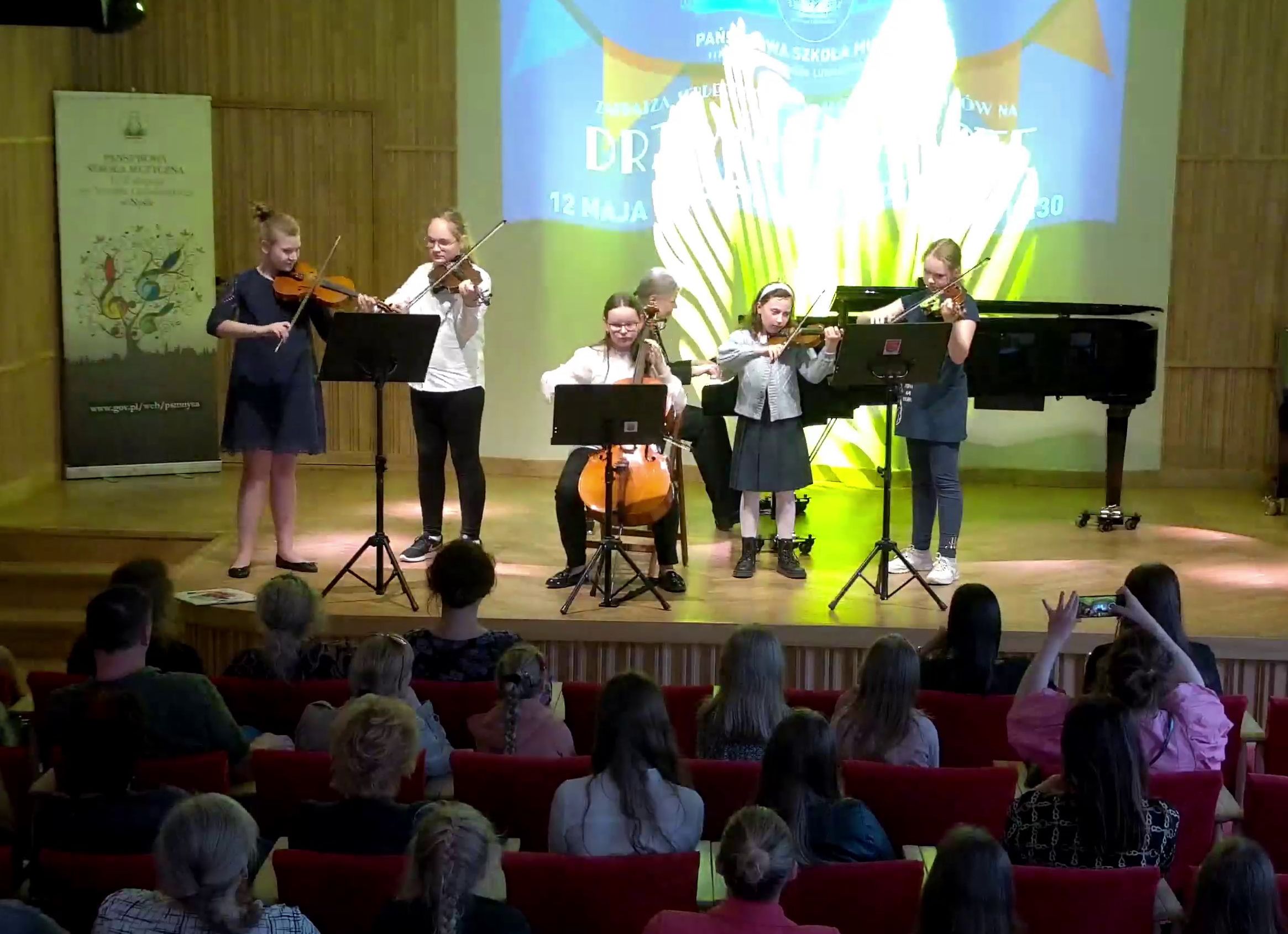 Grafika - zdjęcie. Na estradzie sali koncertowej, przed siedzącą publicznością, zespół smyczkowy z towarzyszeniem fortepianu. W tle wyświetlony jest baner "DRZWI OTWARTE". W lewym rogu z tyłu stoi baner PSM Nysa.